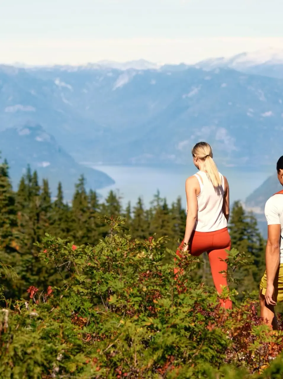 Two hikers walking through dense green forest with mountains and a lake in the background.