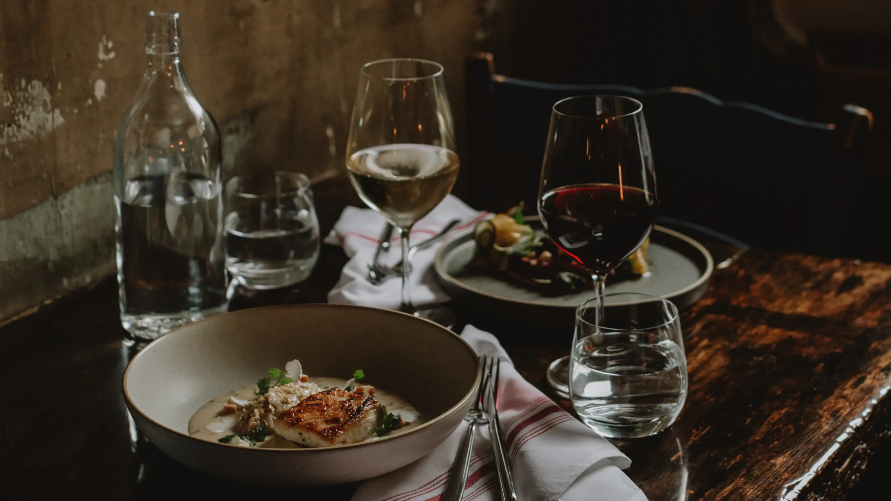 Table with a bowl of creamy soup with grilled fish, two wine glasses with white and red wine, water glasses, and a bottle of water.