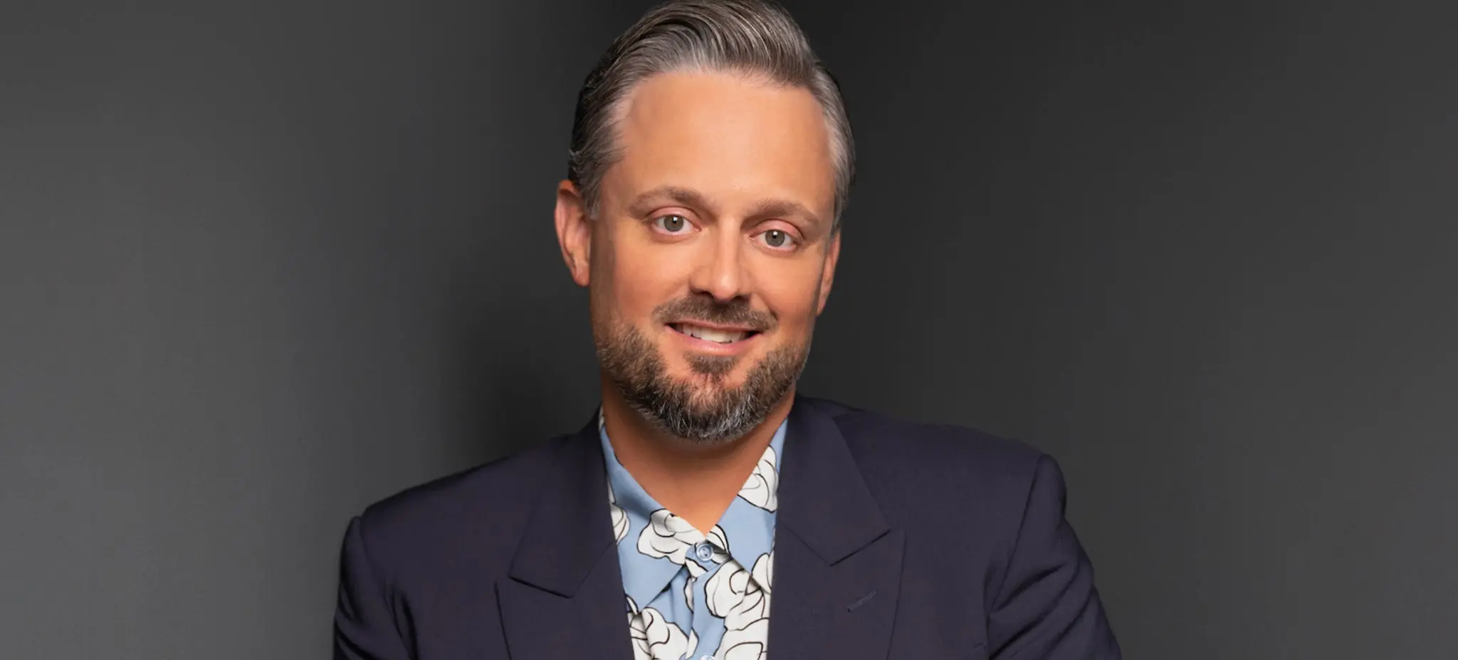 A person with short gray hair and a beard wearing a dark blazer over a blue floral shirt, smiling against a dark background.