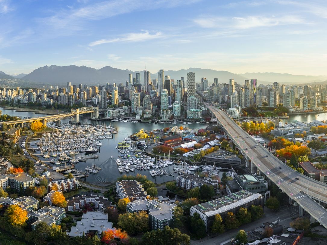 Aerial view of Vancouver's city skyline with high-rise buildings, a marina filled with boats, a bridge, and mountains in the background.