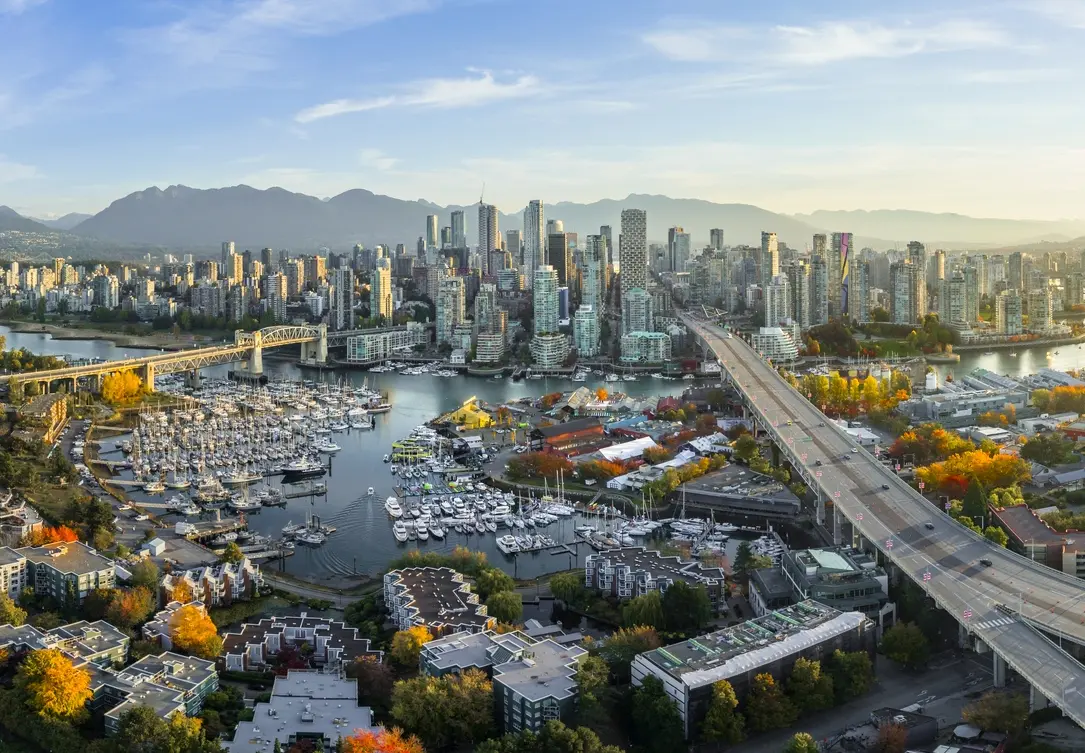 Aerial view of Vancouver's city skyline with high-rise buildings, a marina filled with boats, a bridge, and mountains in the background.