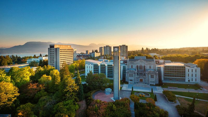 University campus with a tall clock tower, modern and historic buildings, surrounded by trees and mountains in the background at sunset.