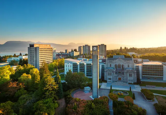 University campus with a tall clock tower, modern and historic buildings, surrounded by trees and mountains in the background at sunset.