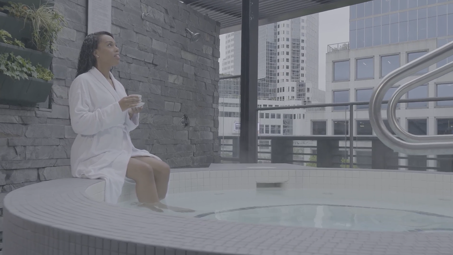 Woman in white bathrobe sitting on edge of hot tub with feet in water, holding cup, with city buildings in background.