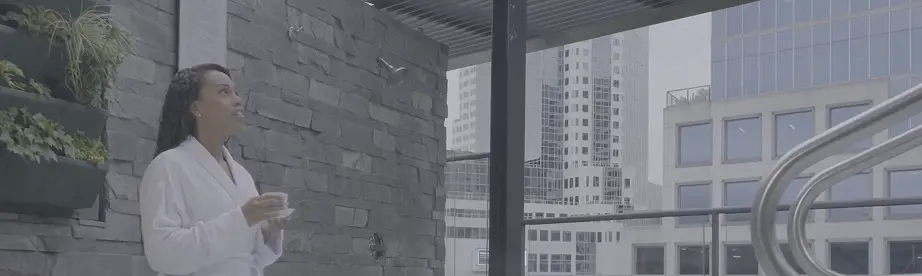 Woman in white bathrobe sitting on edge of hot tub with feet in water, holding cup, with city buildings in background.