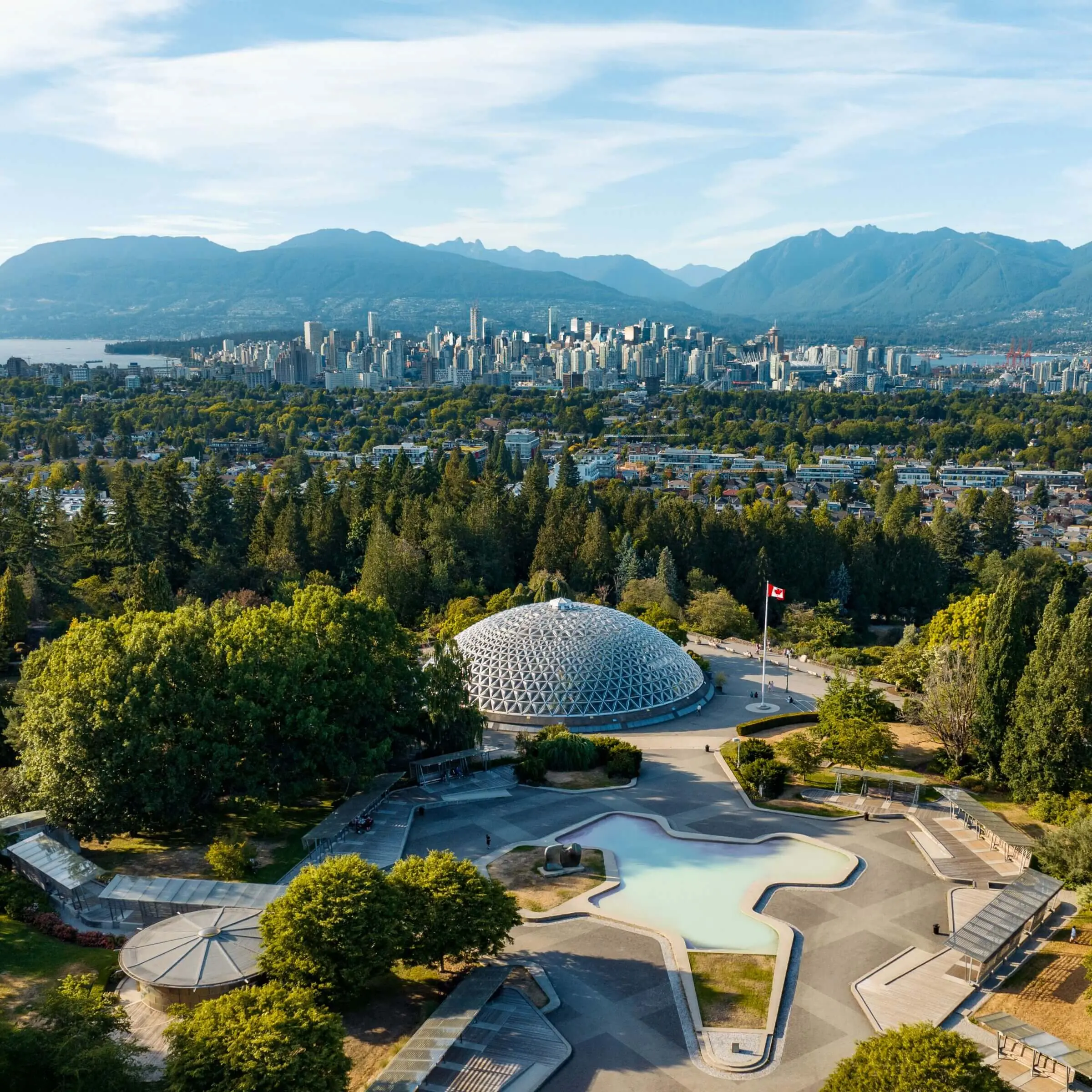 Aerial view of a domed conservatory surrounded by trees, with a winding path and reflecting pool in front, set against a city skyline and mountains—a must-see on any Vancouver itinerary. A Canadian flag is visible near the dome.