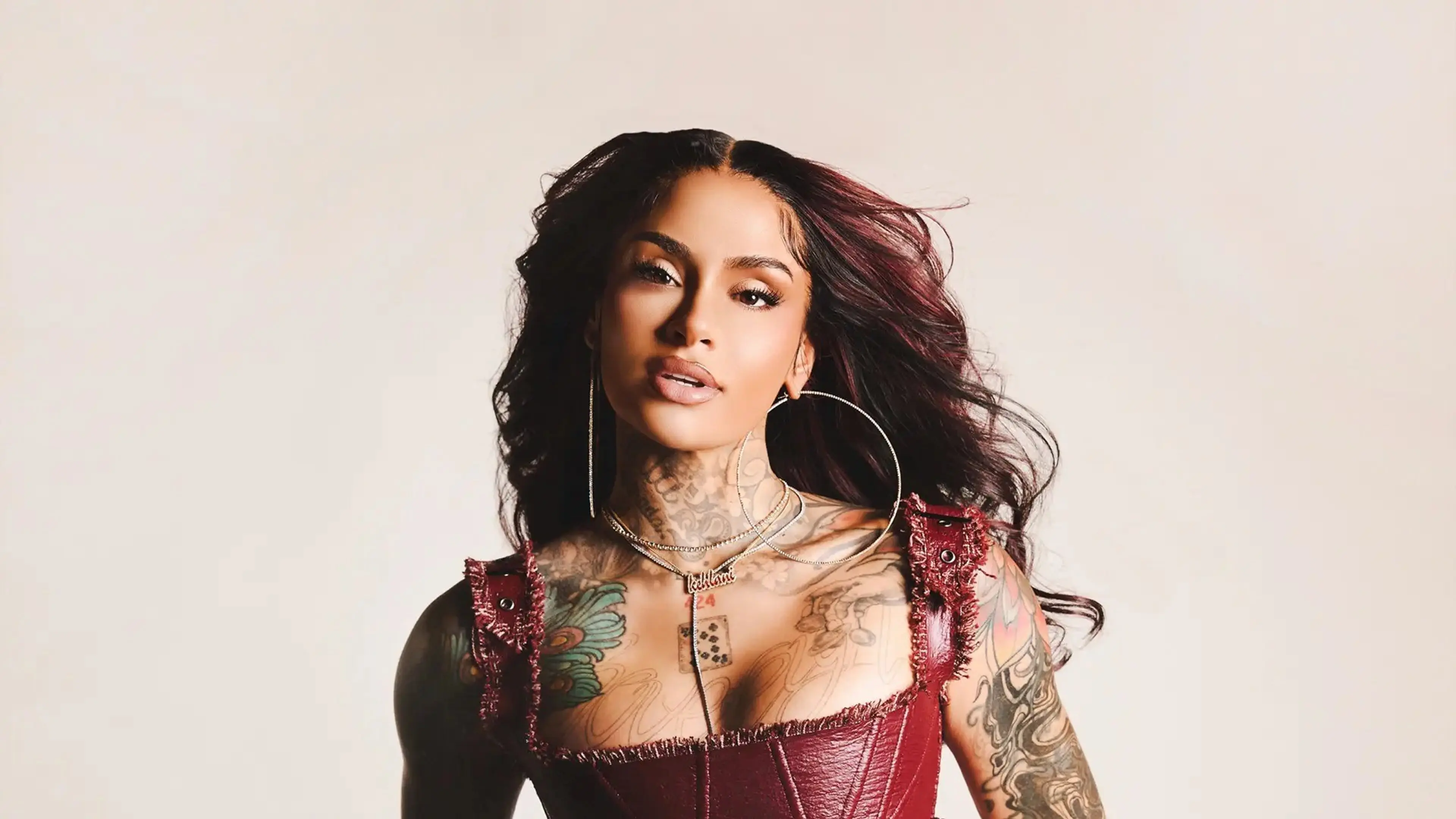 Person with long dark hair wearing large hoop earrings and a red leather top, showing colorful tattoos on arms and chest.