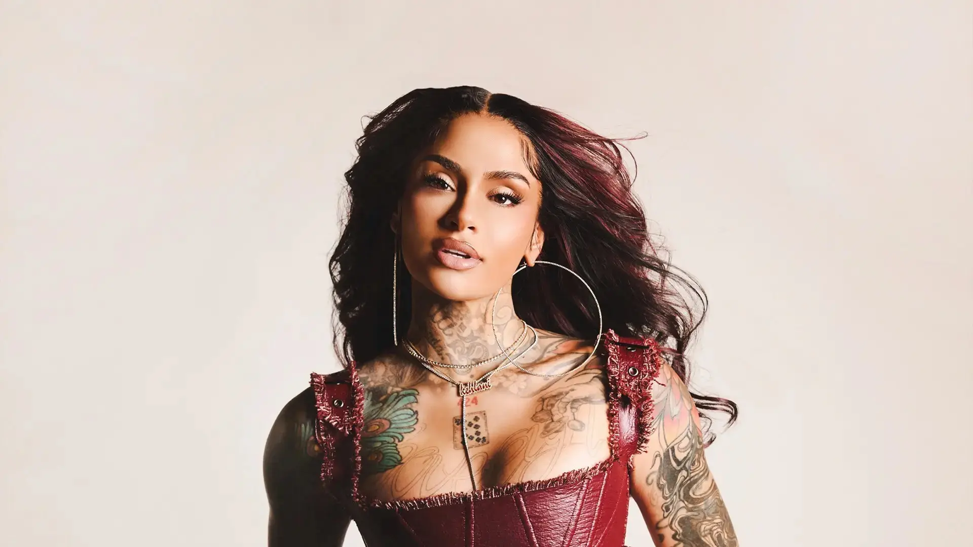 Person with long dark hair wearing large hoop earrings and a red leather top, showing colorful tattoos on arms and chest.