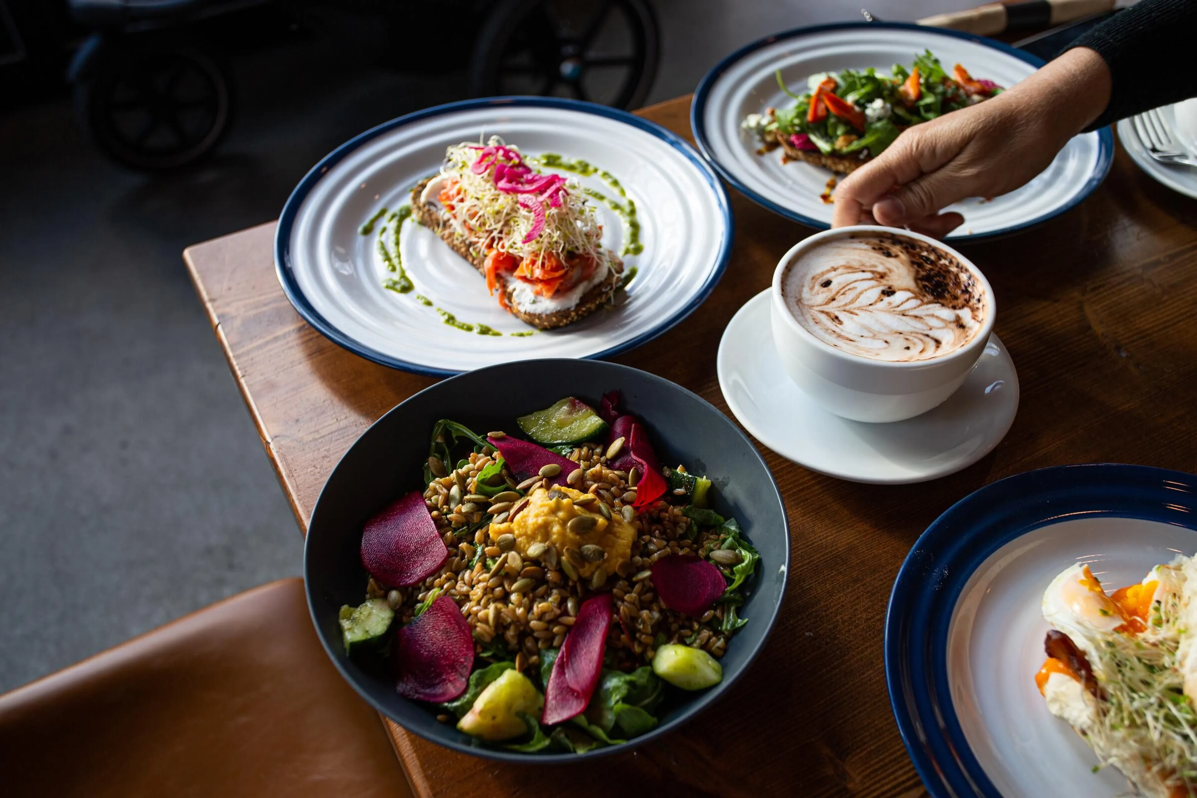 Table with a bowl of salad with grains and vegetables, two plates with open-faced sandwiches, a cup of coffee with latte art, and a hand reaching for the coffee.