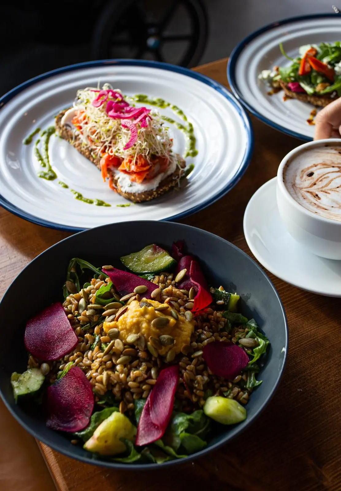 Table with a bowl of salad with grains and vegetables, two plates with open-faced sandwiches, a cup of coffee with latte art, and a hand reaching for the coffee.