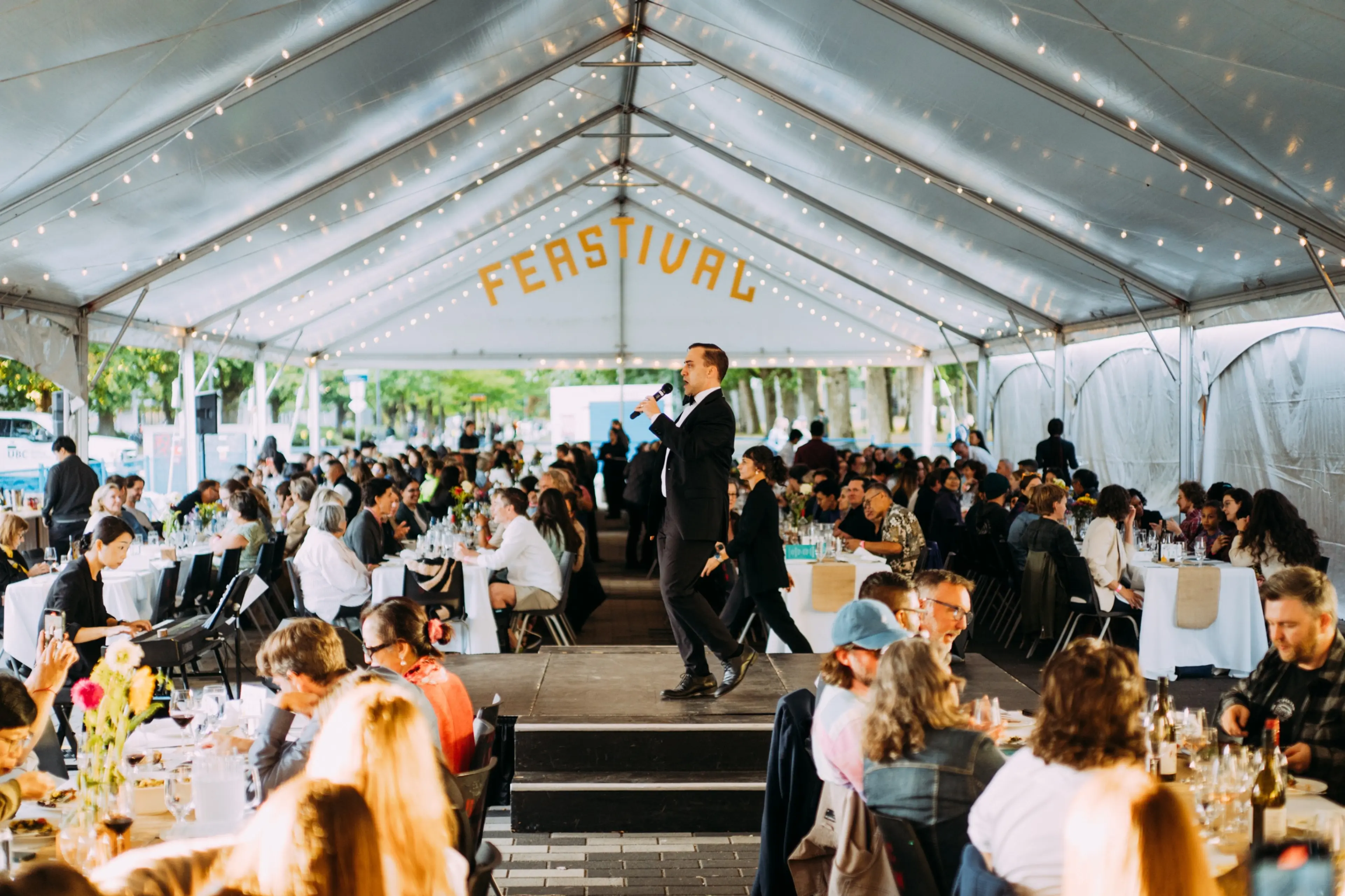 Person in a suit speaking into a microphone on a stage under a tent with many people seated at tables for a Feastival event.