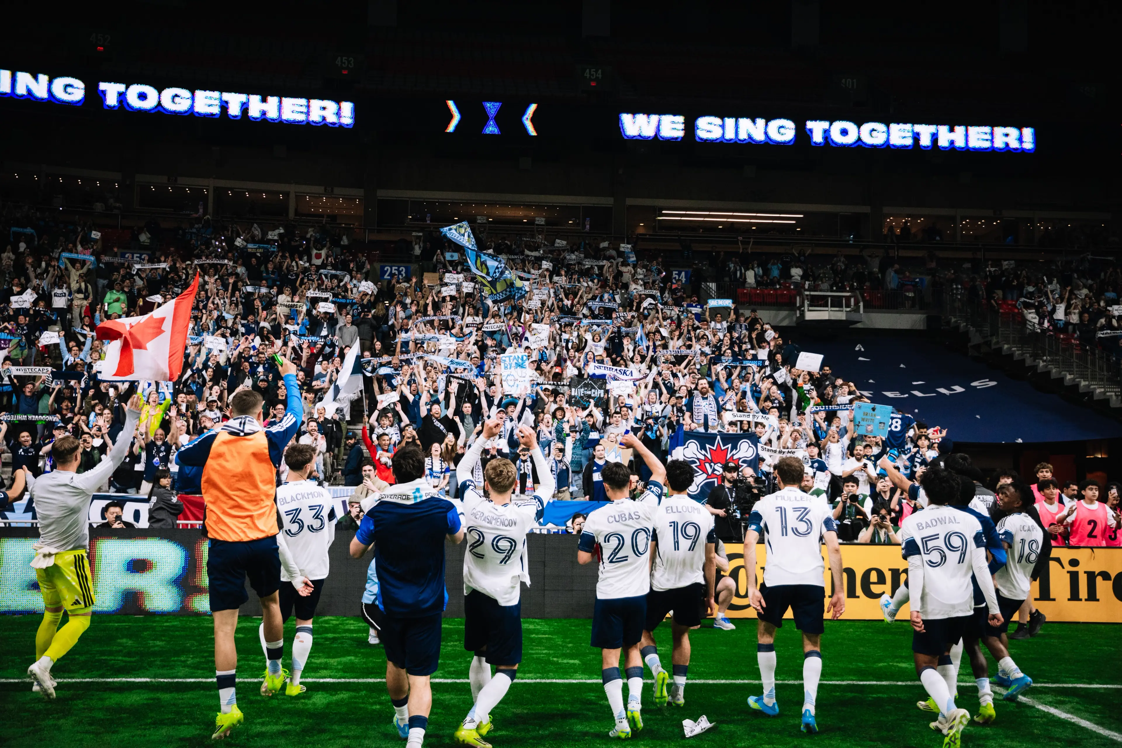 Soccer players in white jerseys facing a cheering crowd with raised arms and flags in a stadium.