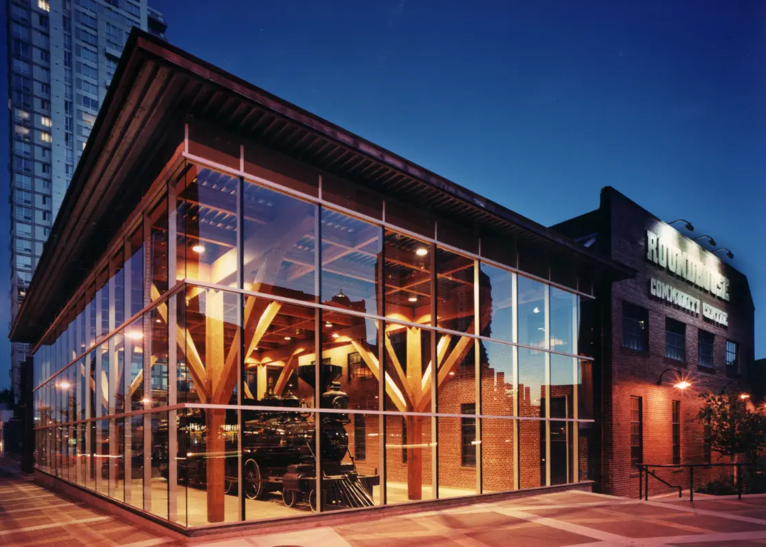 Glass building with wooden beams displaying a vintage steam locomotive inside at dusk.