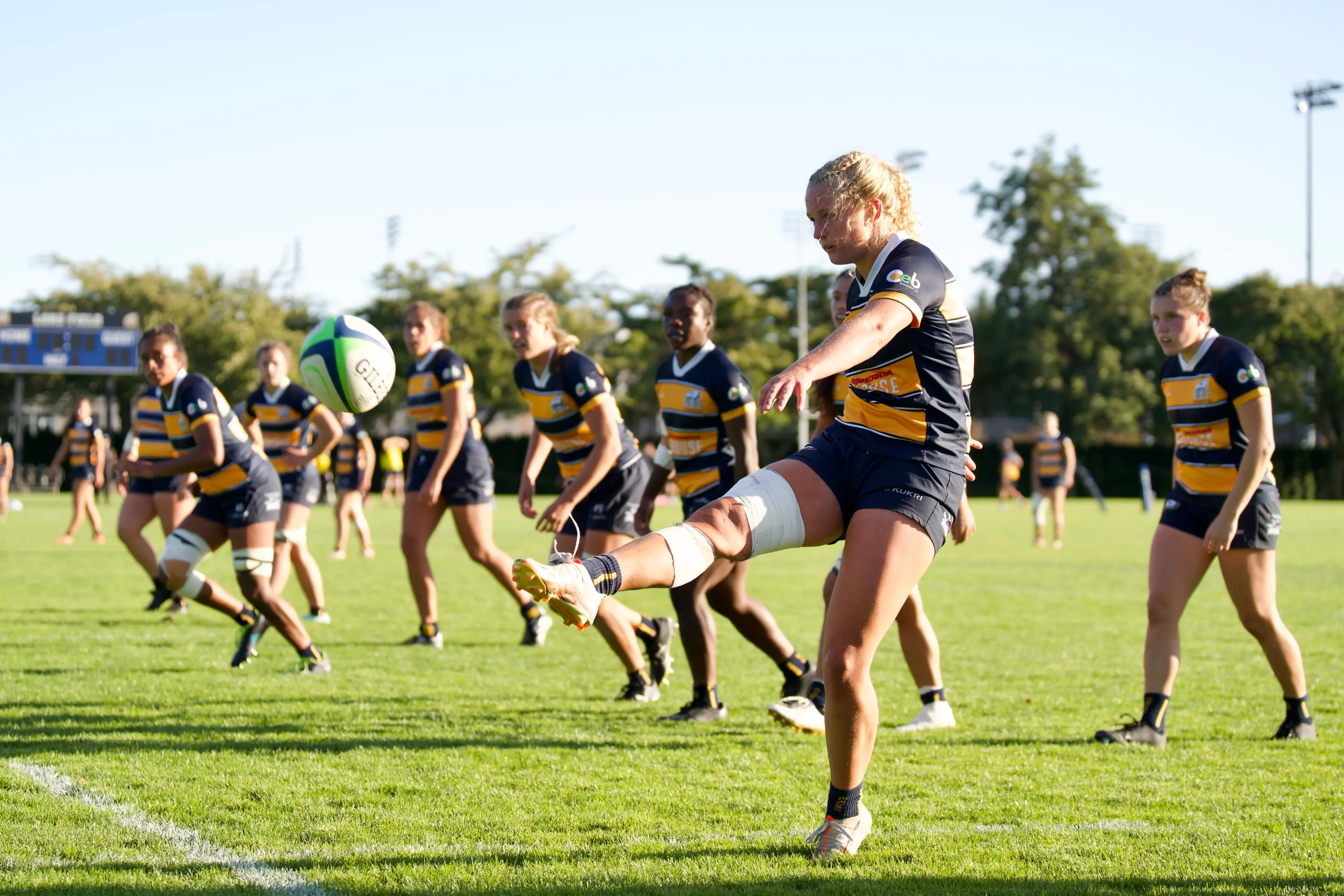 A team of women rugby players on the field with one person kicking the ball during a match.