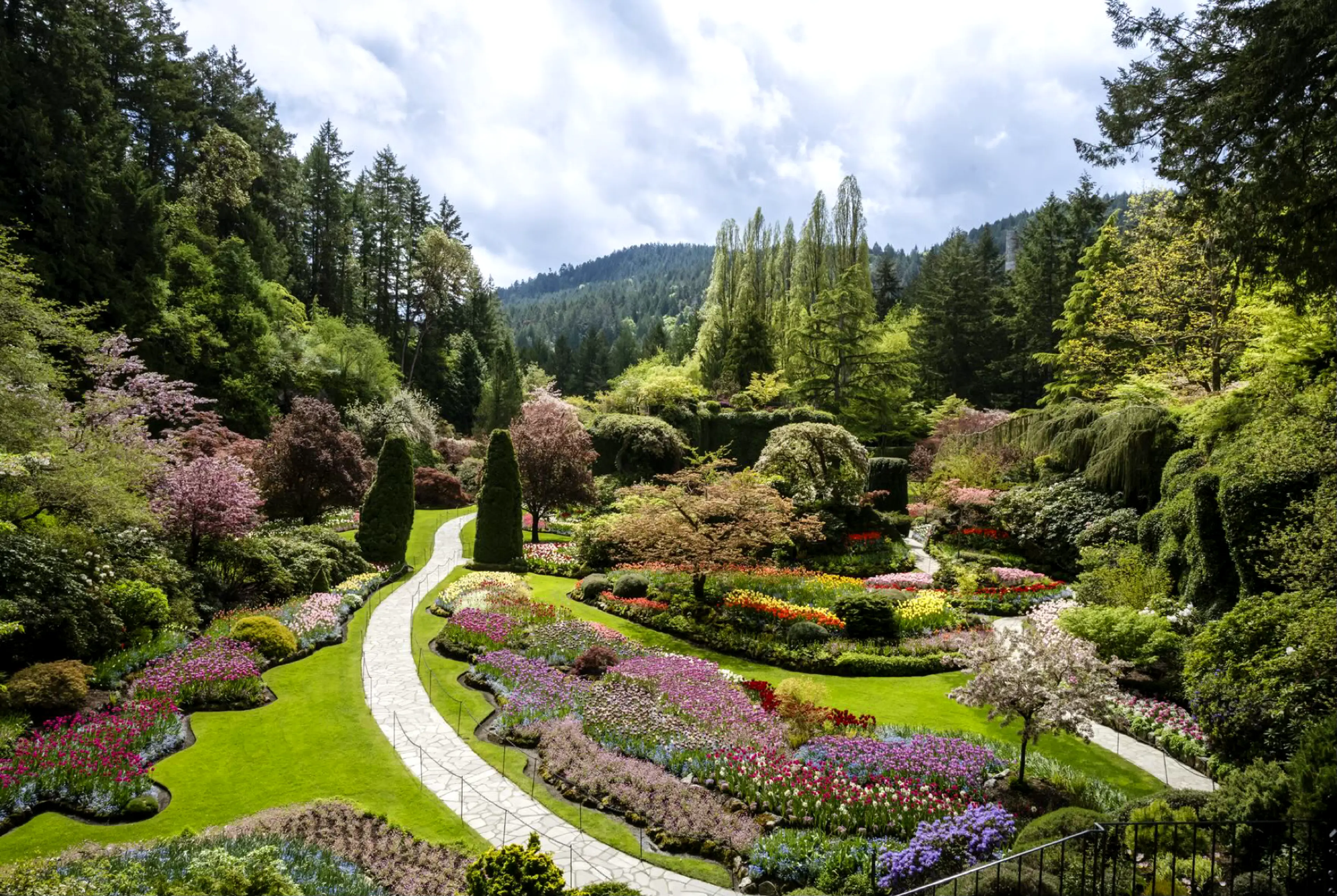 A vibrant botanical garden with winding stone paths, colorful flower beds, manicured lawns, and lush green trees—an ideal spot for day trips from Vancouver—set against forested hills under a partly cloudy sky.