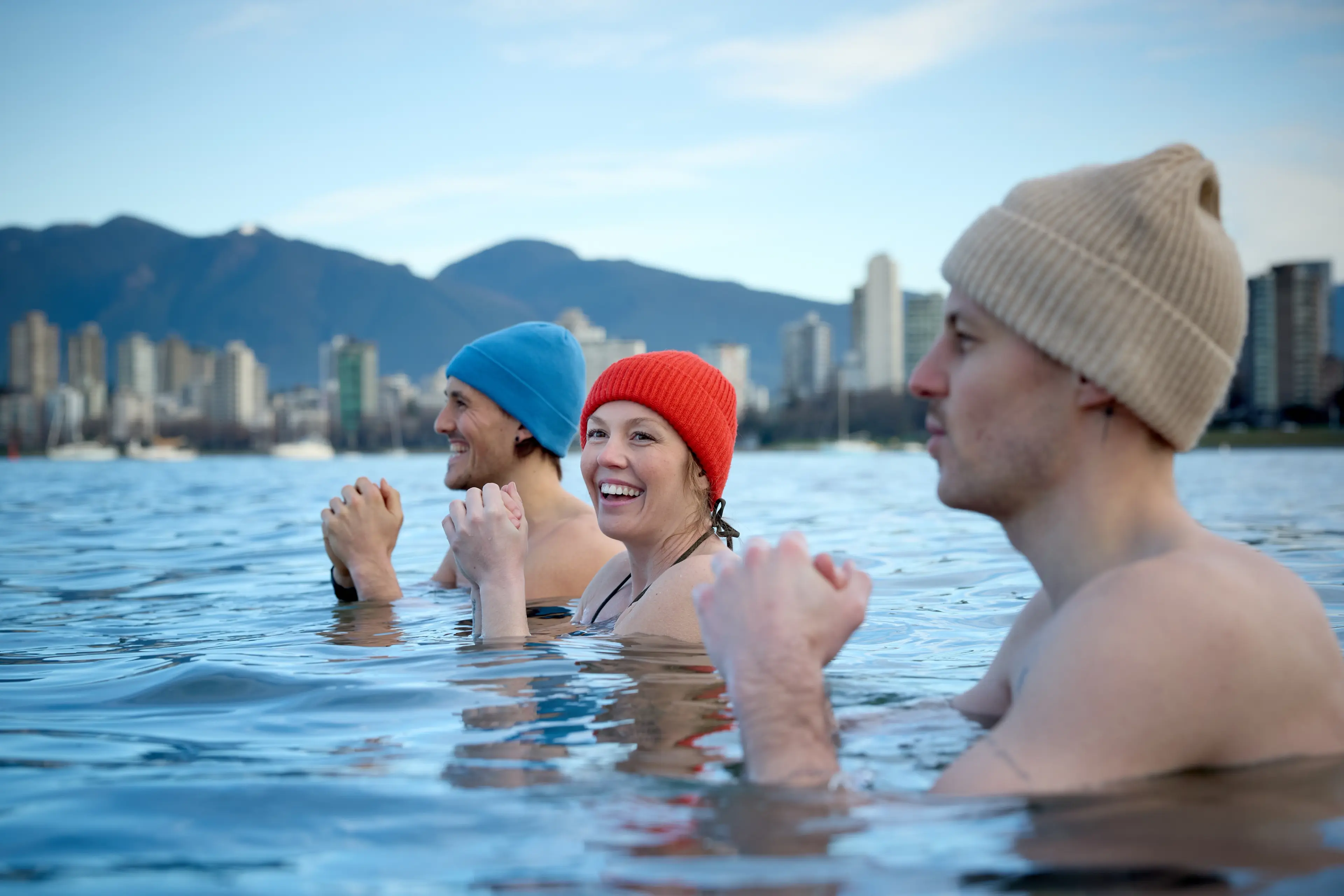 Three people wearing colorful knit caps standing chest-deep in water with a city skyline and mountains in the background.