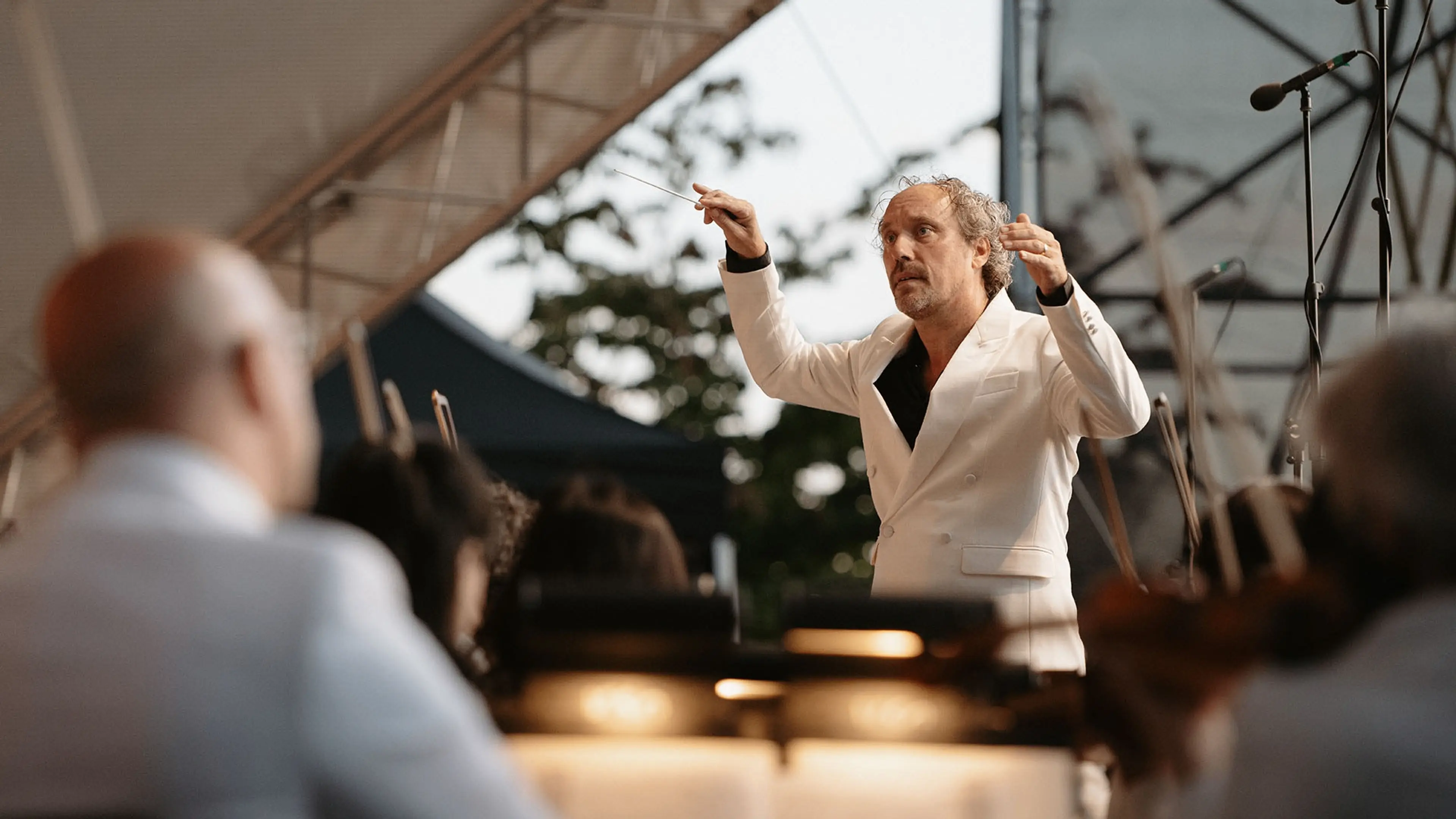 A conductor in a white suit leads an orchestra during an outdoor performance under a tent.