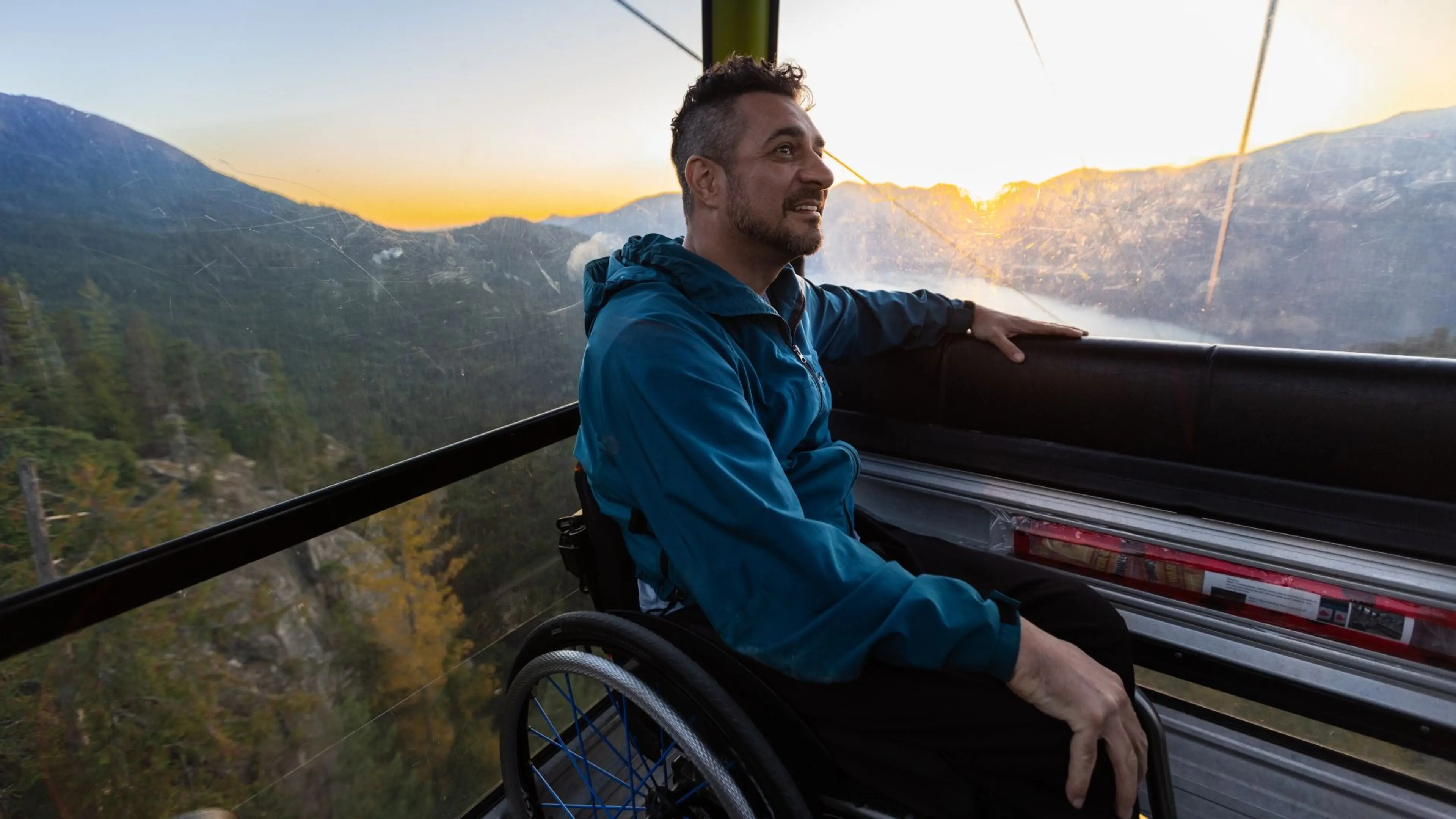 A man in a manual wheelchair sits comfortably inside a glass-enclosed gondola cabin, looking out at a stunning mountain sunset.