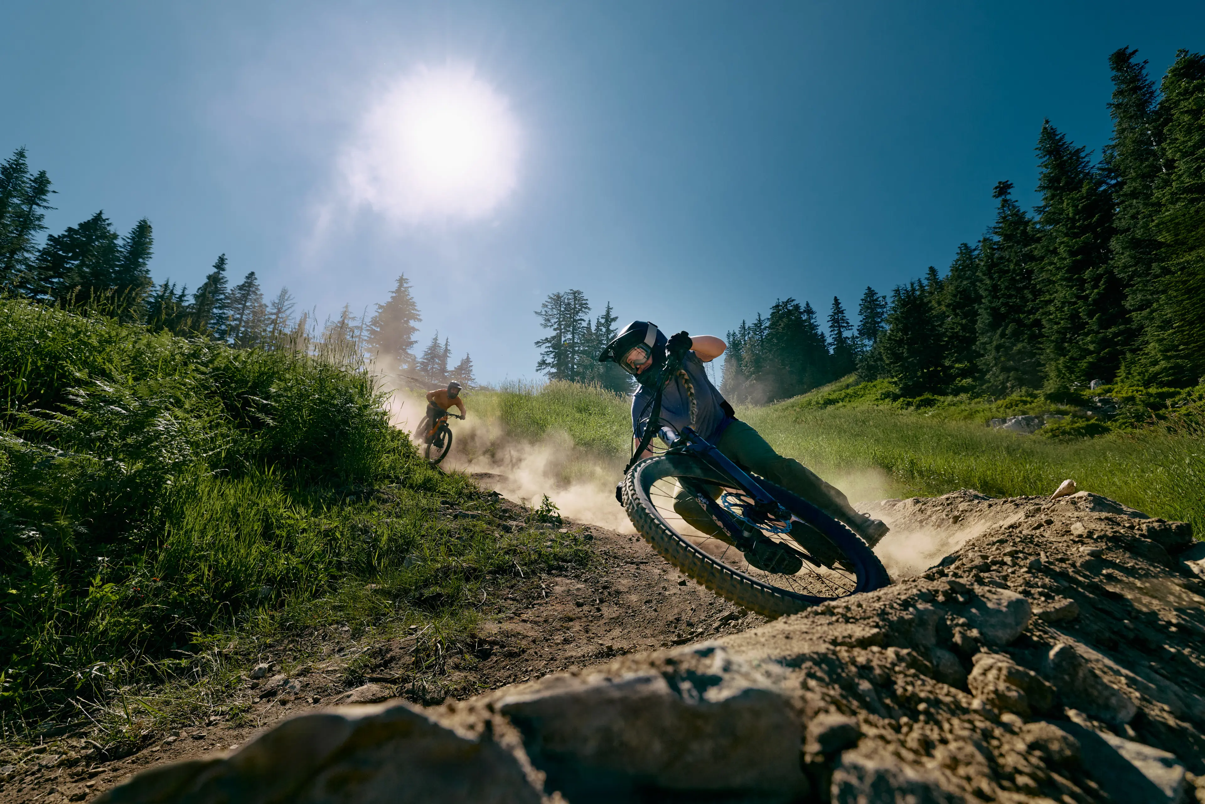 Two mountain bikers wearing helmets riding on a dirt trail surrounded by green grass and trees under a bright sun.