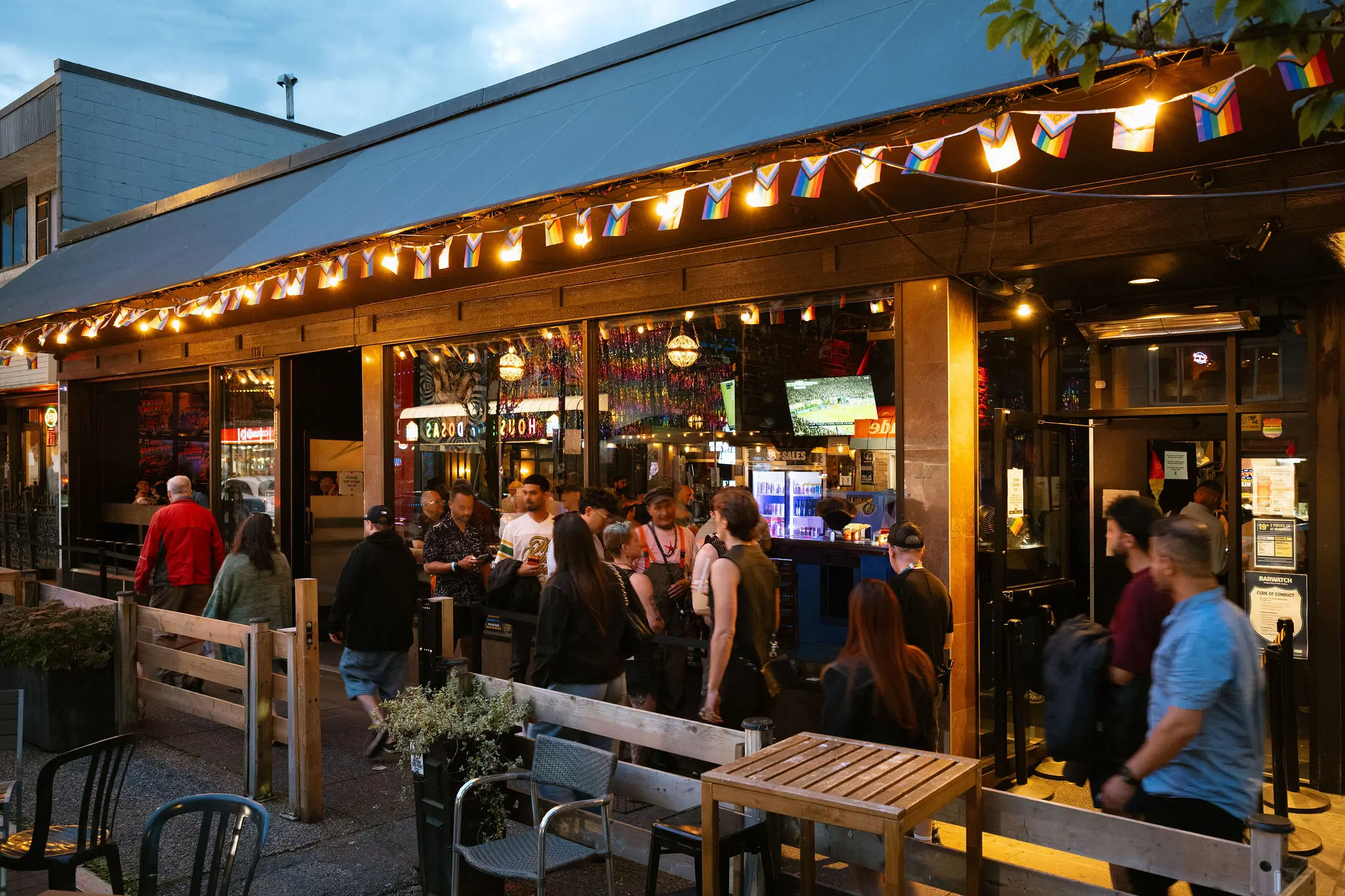 Crowd of people outside a bar with string lights and small rainbow flags hanging from the roof edge during evening.