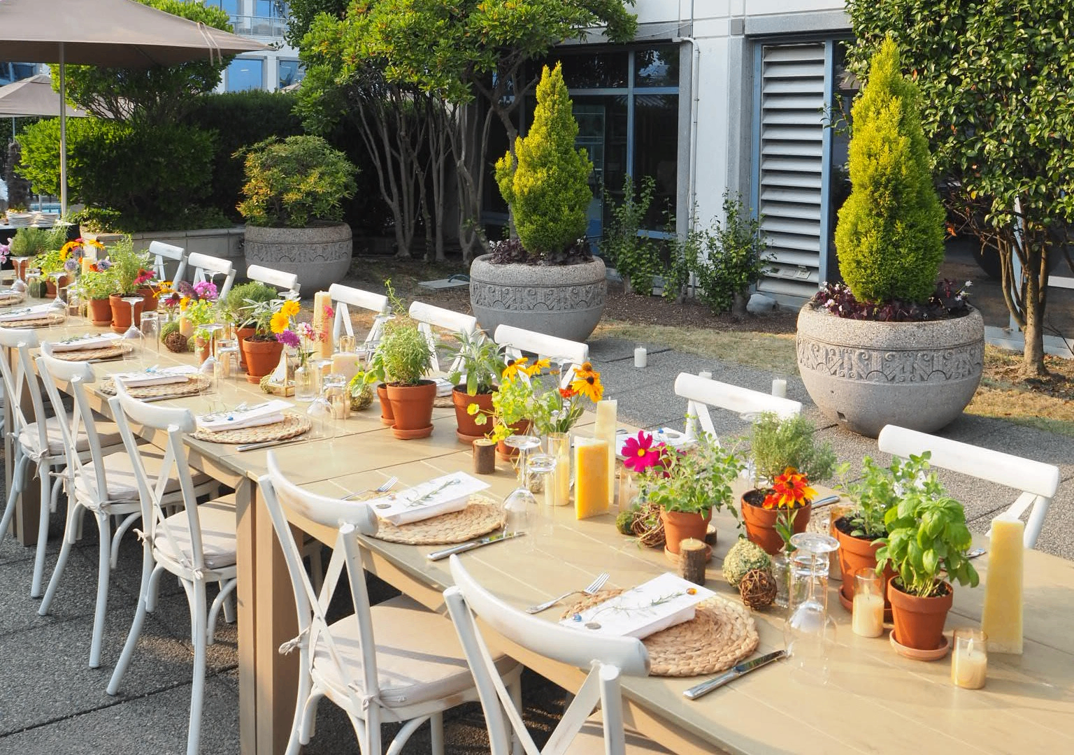Long outdoor dining table with white chairs, decorated with potted plants, flowers, candles, and place settings.