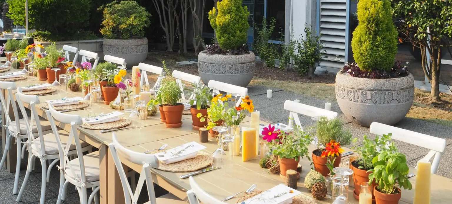 Long outdoor dining table with white chairs, decorated with potted plants, flowers, candles, and place settings.