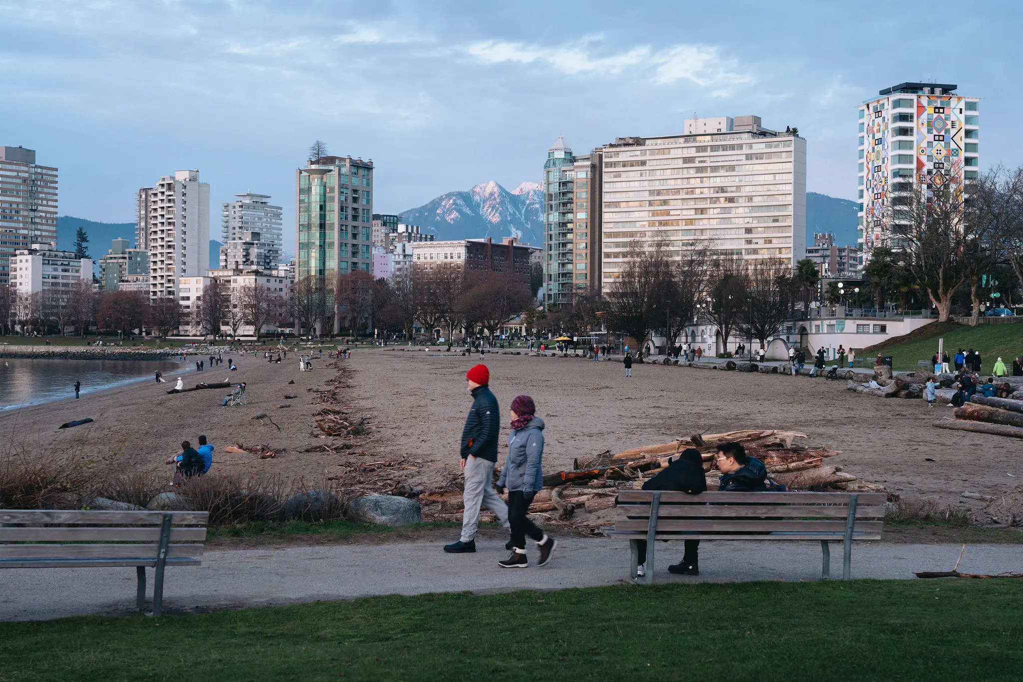 People walking and sitting on benches near a sandy beach with city buildings and mountains in the background.