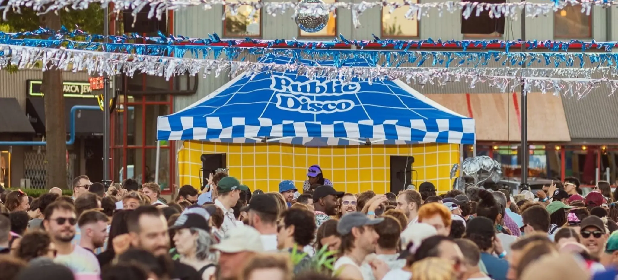 Crowd of people at an outdoor event in front of a blue and white tent labeled Public Disco with blue and white streamers overhead.
