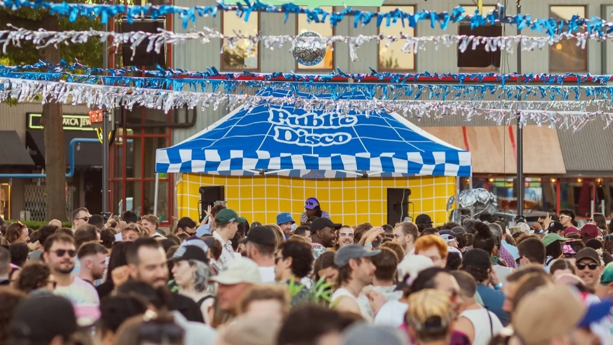 Crowd of people at an outdoor event in front of a blue and white tent labeled Public Disco with blue and white streamers overhead.
