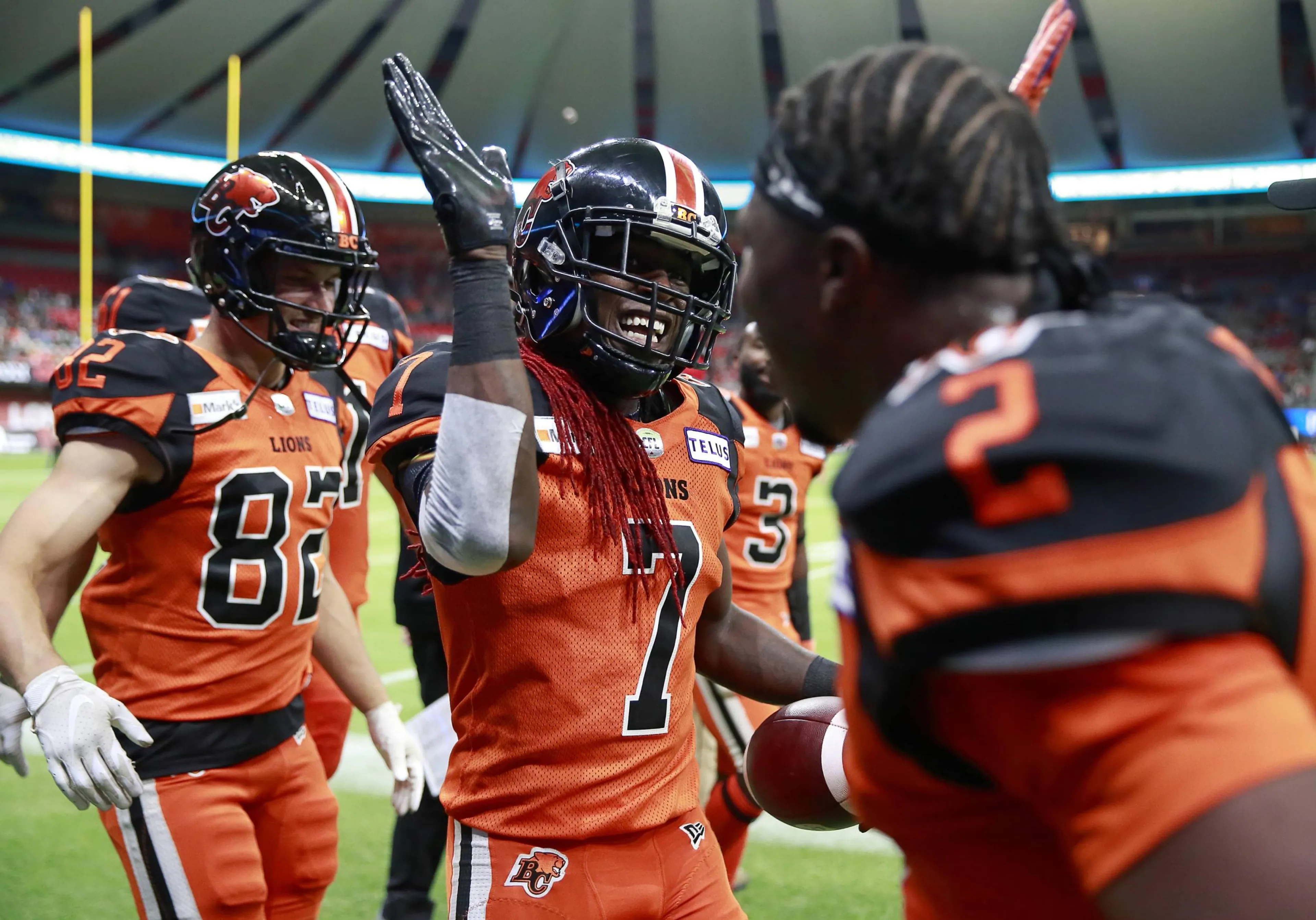 Football players in orange and black BC Lions uniforms celebrating on the field, one player holding a football and raising his hand for a high-five.