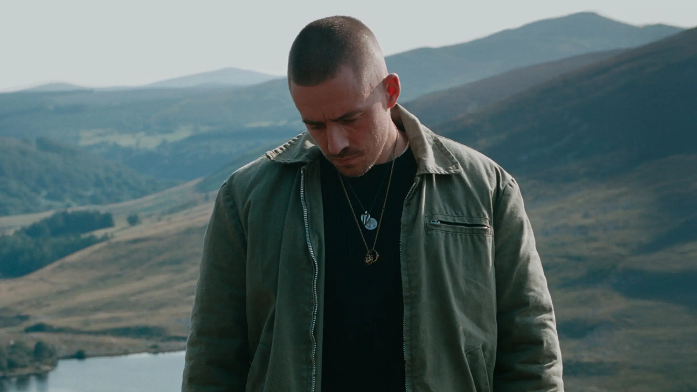 A person with short hair and a mustache wearing a green jacket and black shirt stands outdoors with mountains and a lake in the background.
