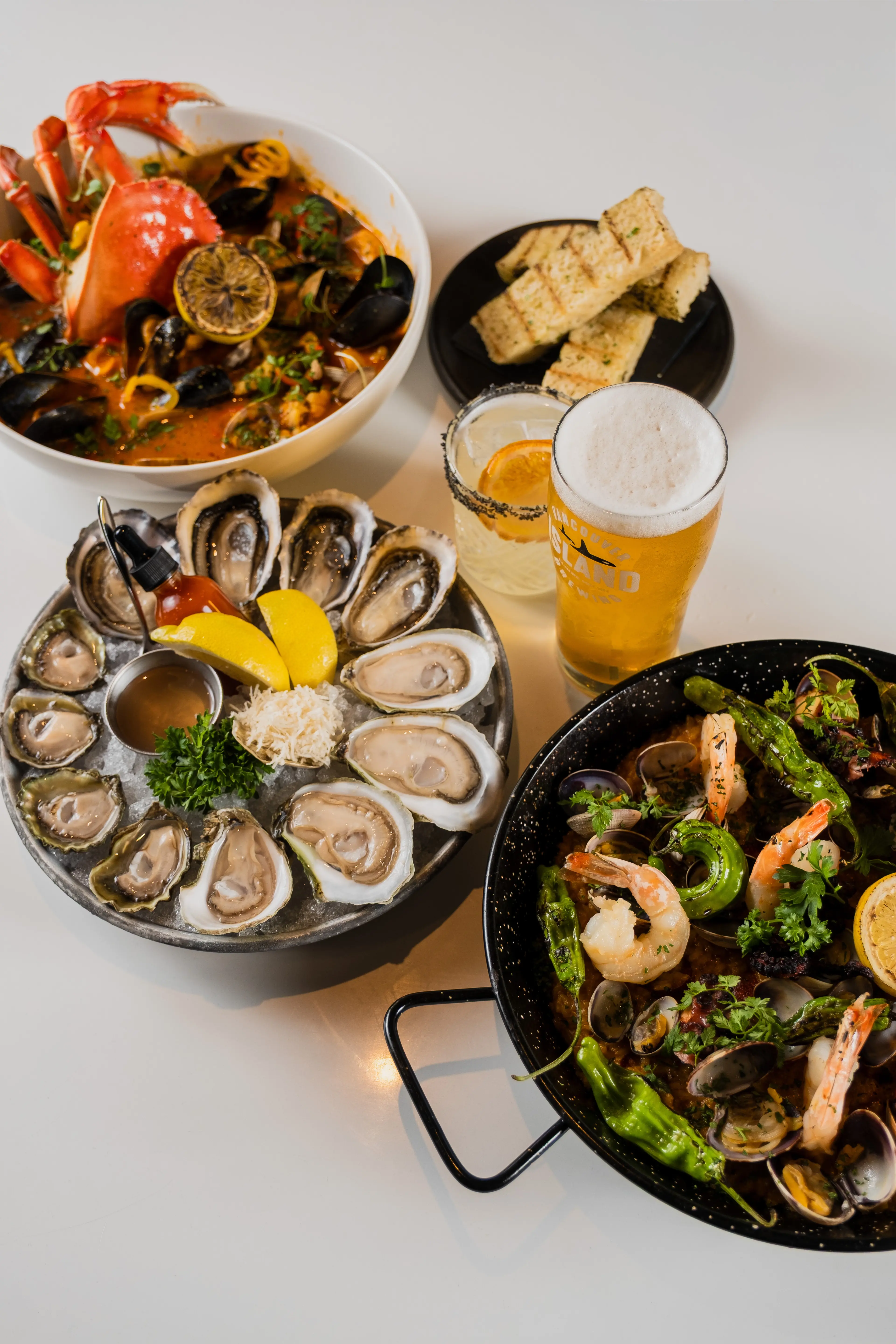Seafood dishes including oysters on ice with lemon wedges, a bowl with crab and mussels, a pan with shrimp and clams, grilled bread, a glass of beer, and a cocktail with an orange slice.