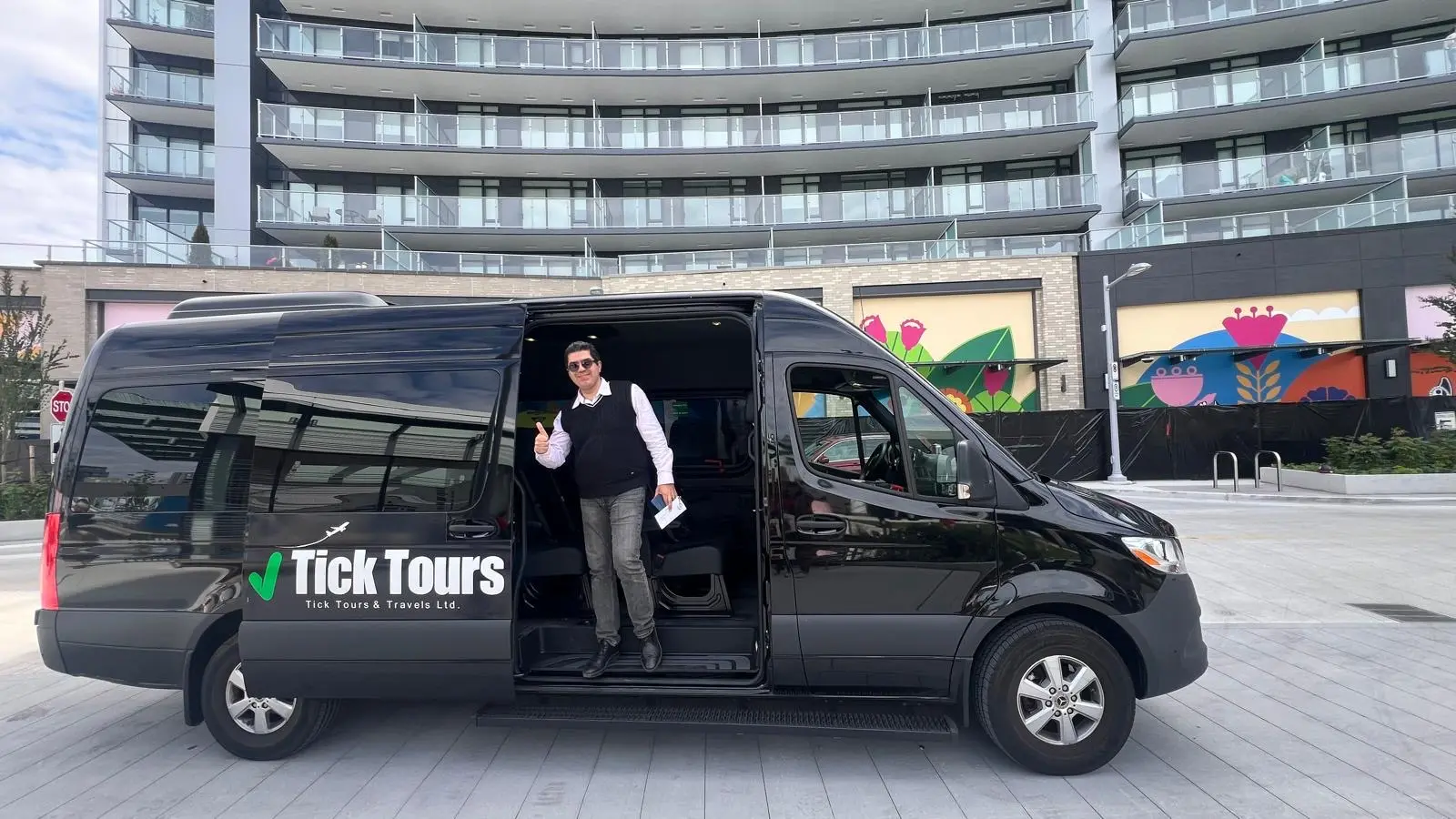 Man in sunglasses giving thumbs up standing in open door of black Tick Tours van in urban area.