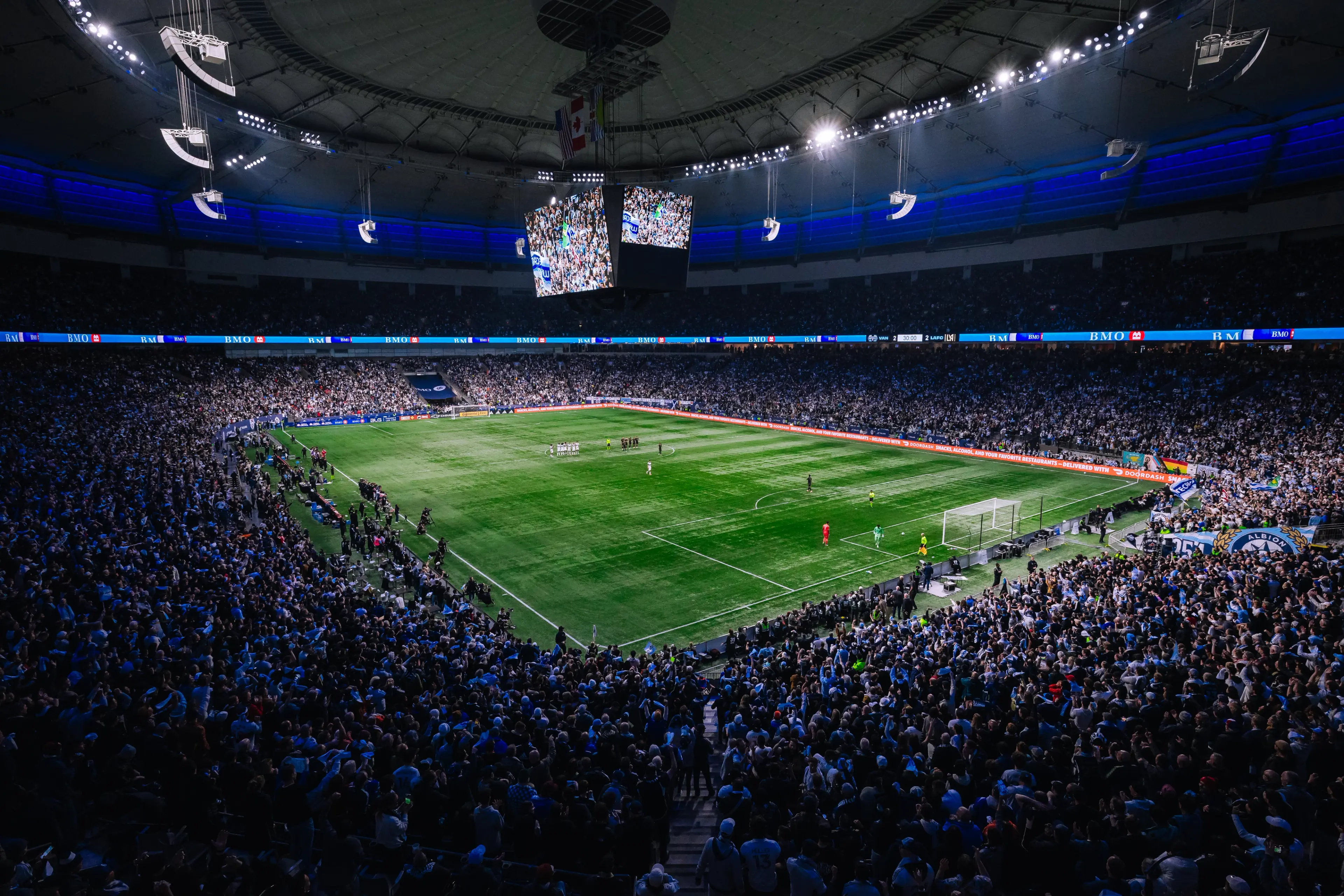 A packed stadium with a soccer field in the center, players on the field, and a large screen hanging from the ceiling.