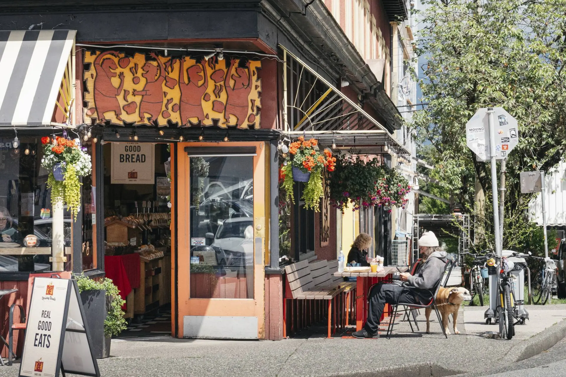 Outdoor seating at a bakery with a sign reading Good Bread and Real Good Eats, two people sitting at a table, and a dog beside one person.