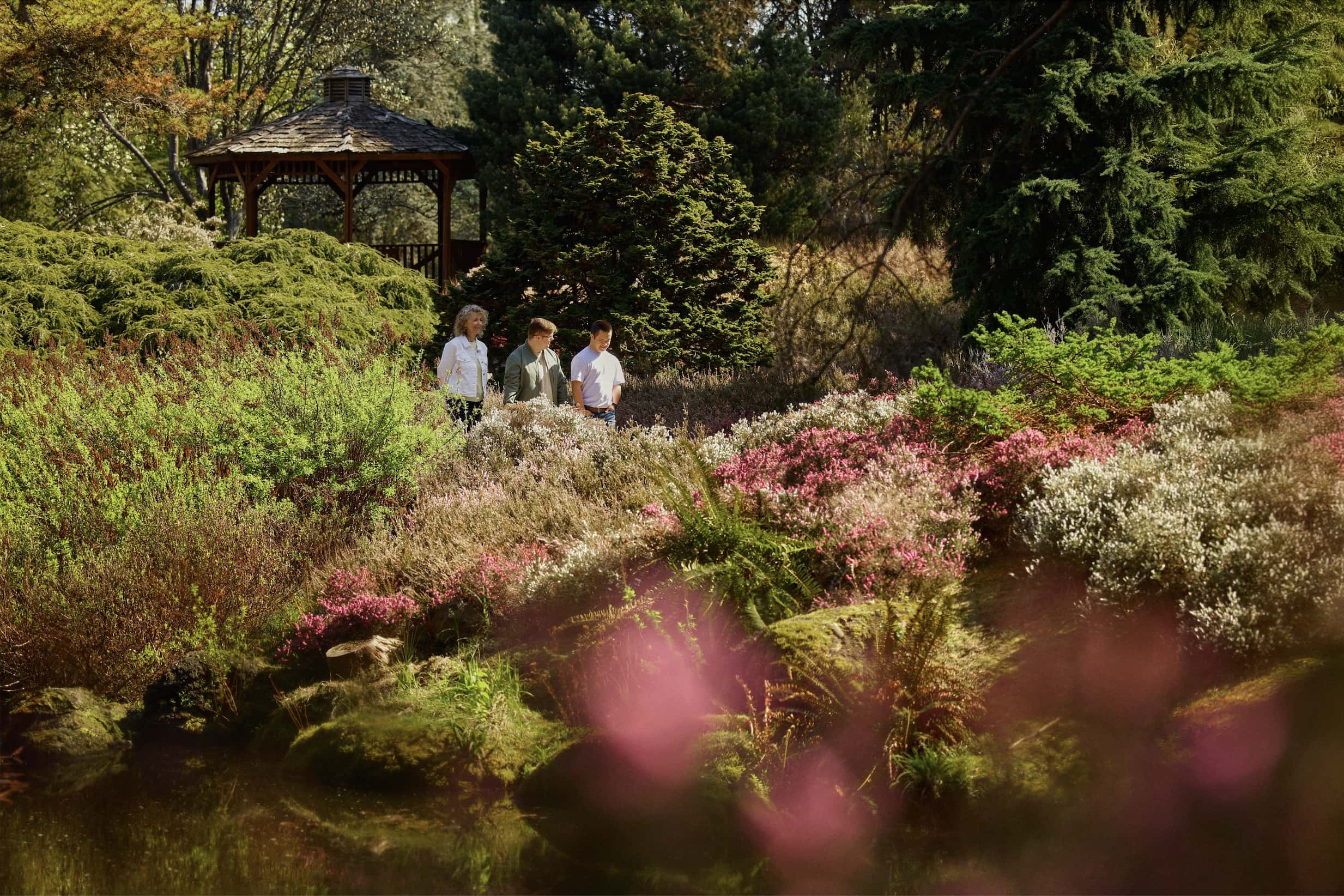 Three people walk through a vibrant garden with a gazebo, surrounded by colorful flowering plants and lush greenery under sunny skies.