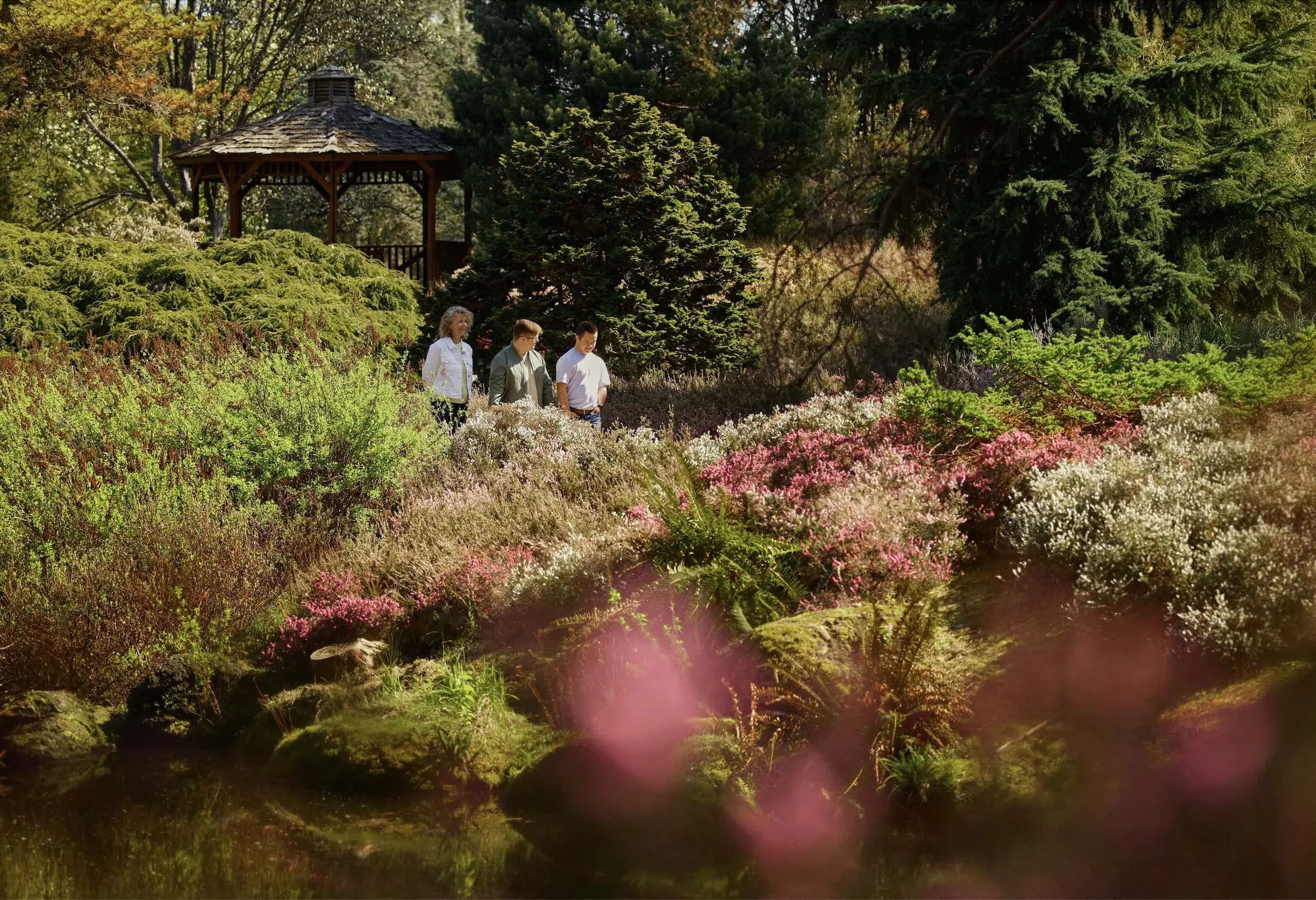 Three people walk through a vibrant garden with a gazebo, surrounded by colorful flowering plants and lush greenery under sunny skies.