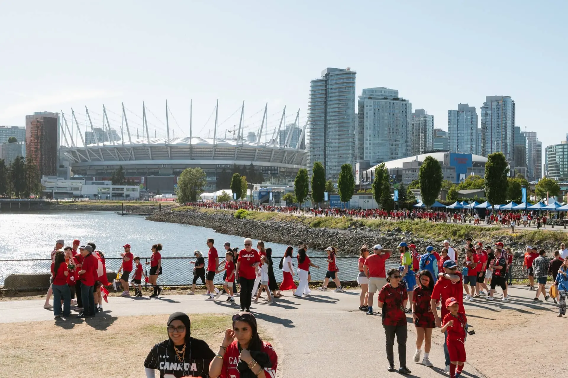 Crowd of people wearing red near waterfront with stadium and city skyline in the background on a sunny day.