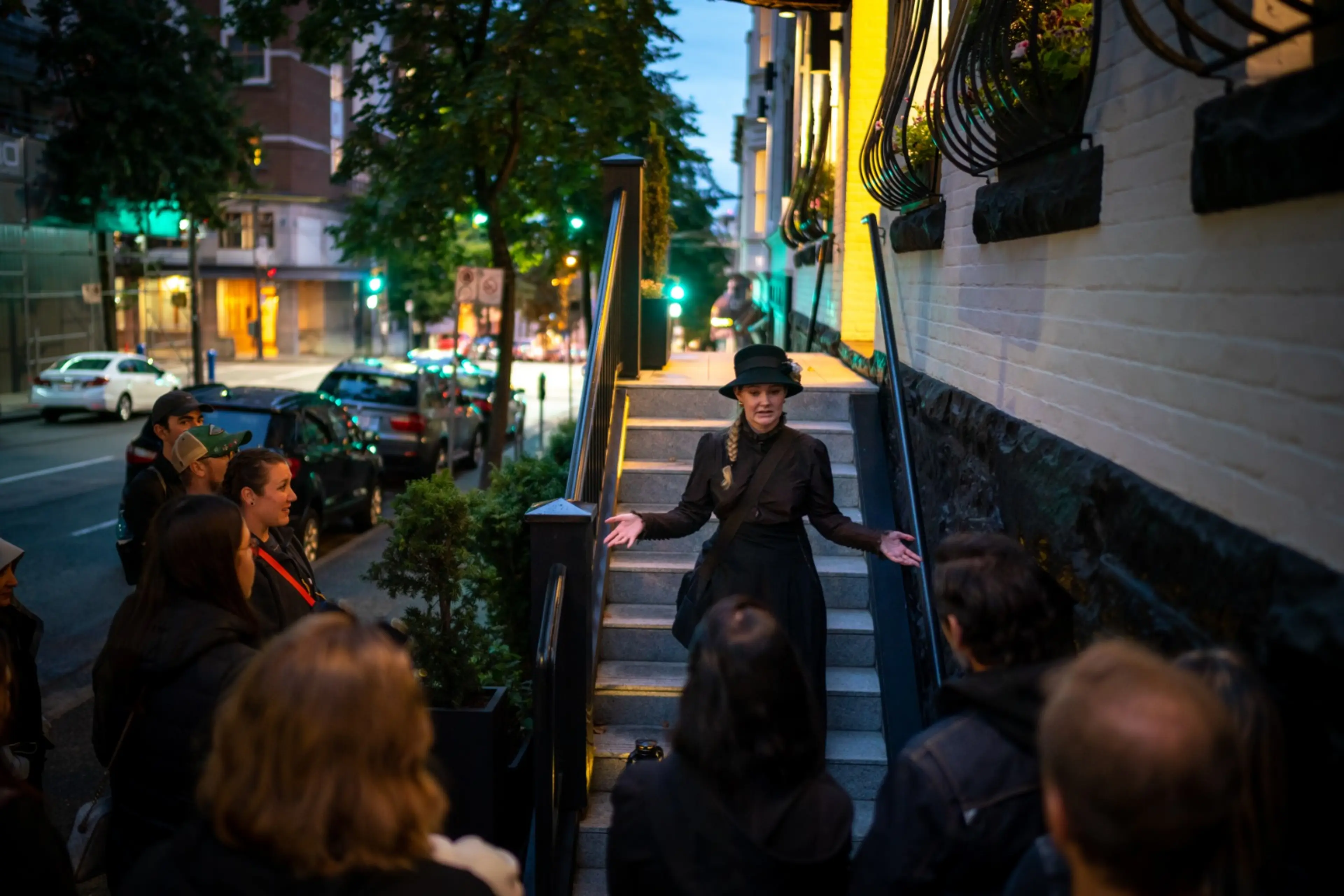 Woman in black dress and hat speaking to a group on a city sidewalk near stairs at dusk.