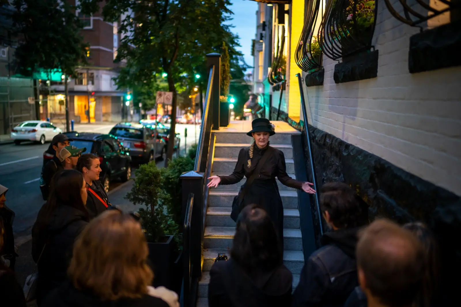 Woman in black dress and hat speaking to a group on a city sidewalk near stairs at dusk.