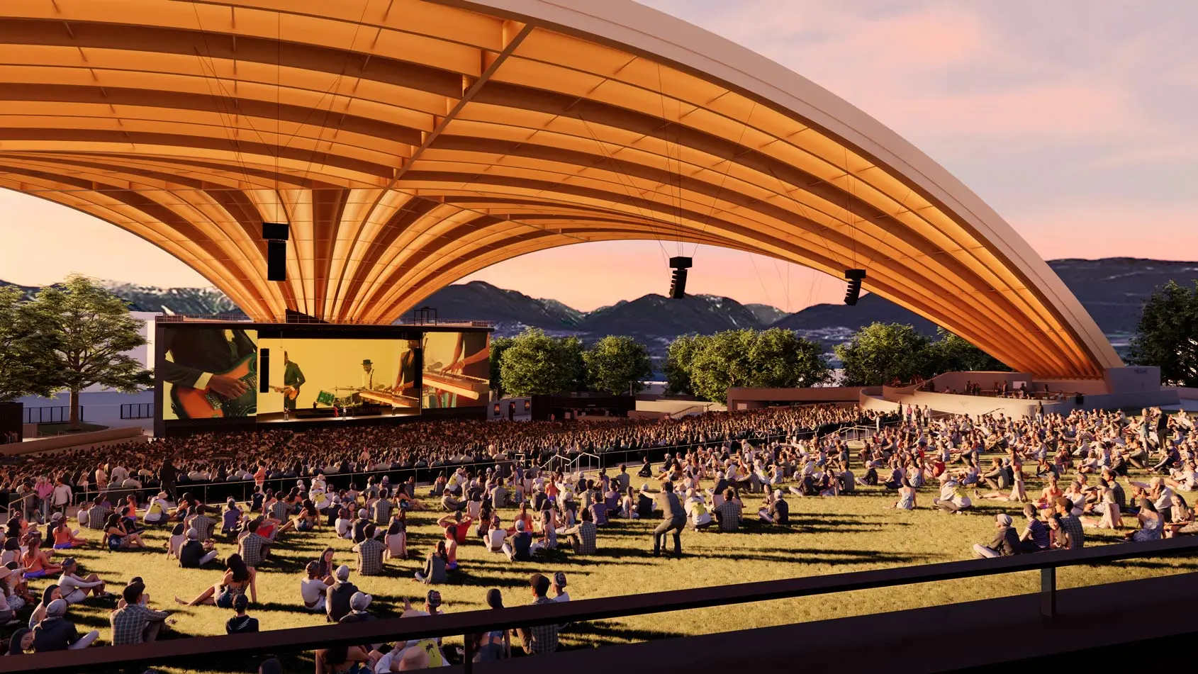 Large crowd seated on grass under a wooden arched pavilion watching a live band on stage with mountains in the background at sunset.
