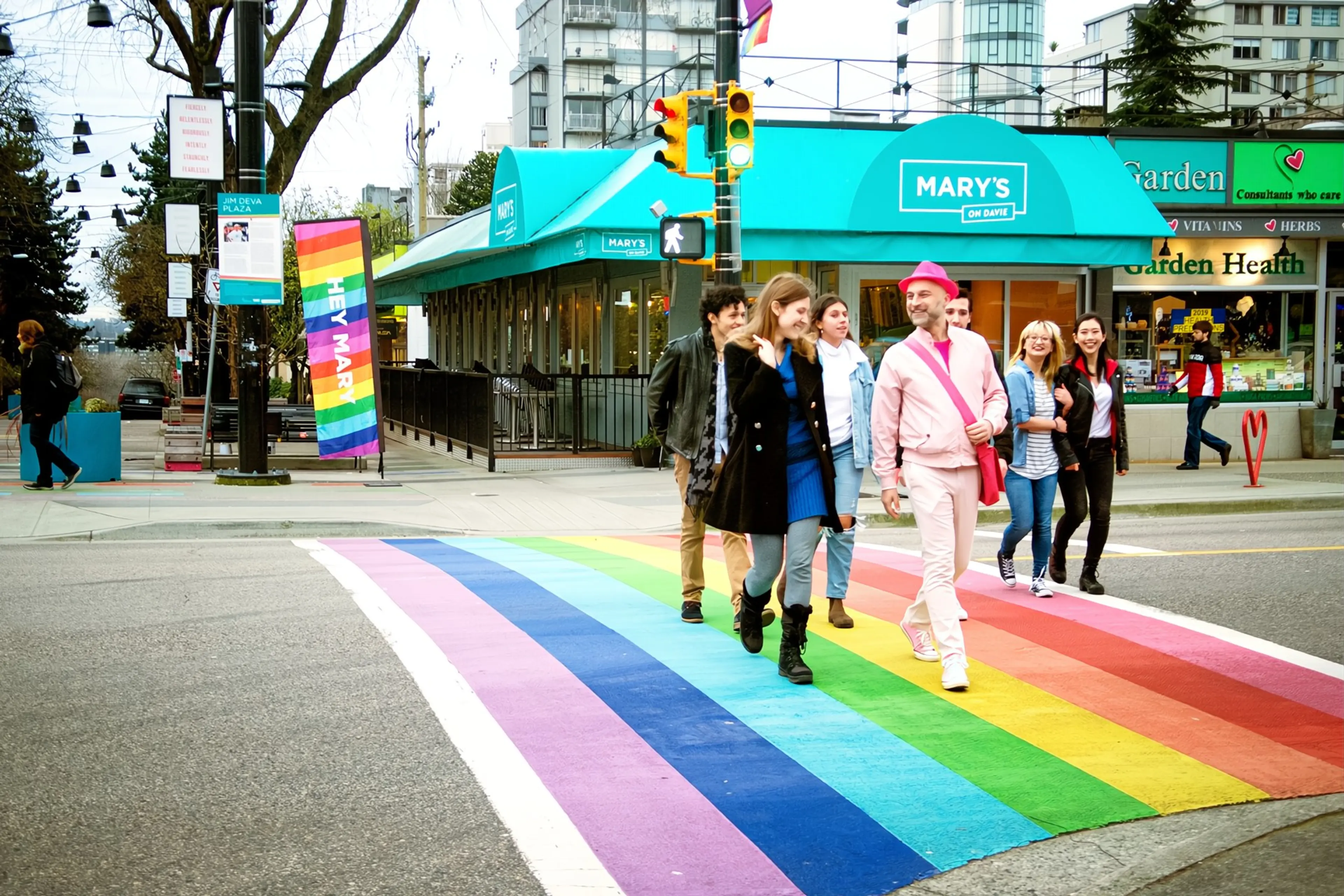 Six people walking across a rainbow-colored crosswalk near a teal awning with the sign 'Mary's on Davie' in an urban setting.
