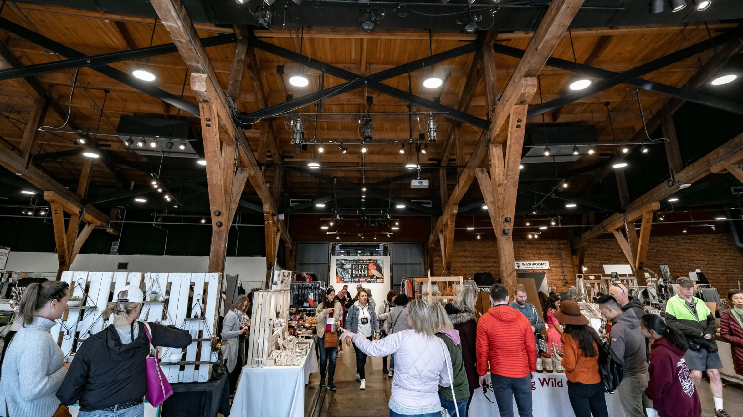 Indoor market with wooden beams and ceiling, people browsing various vendor tables with crafts and goods.