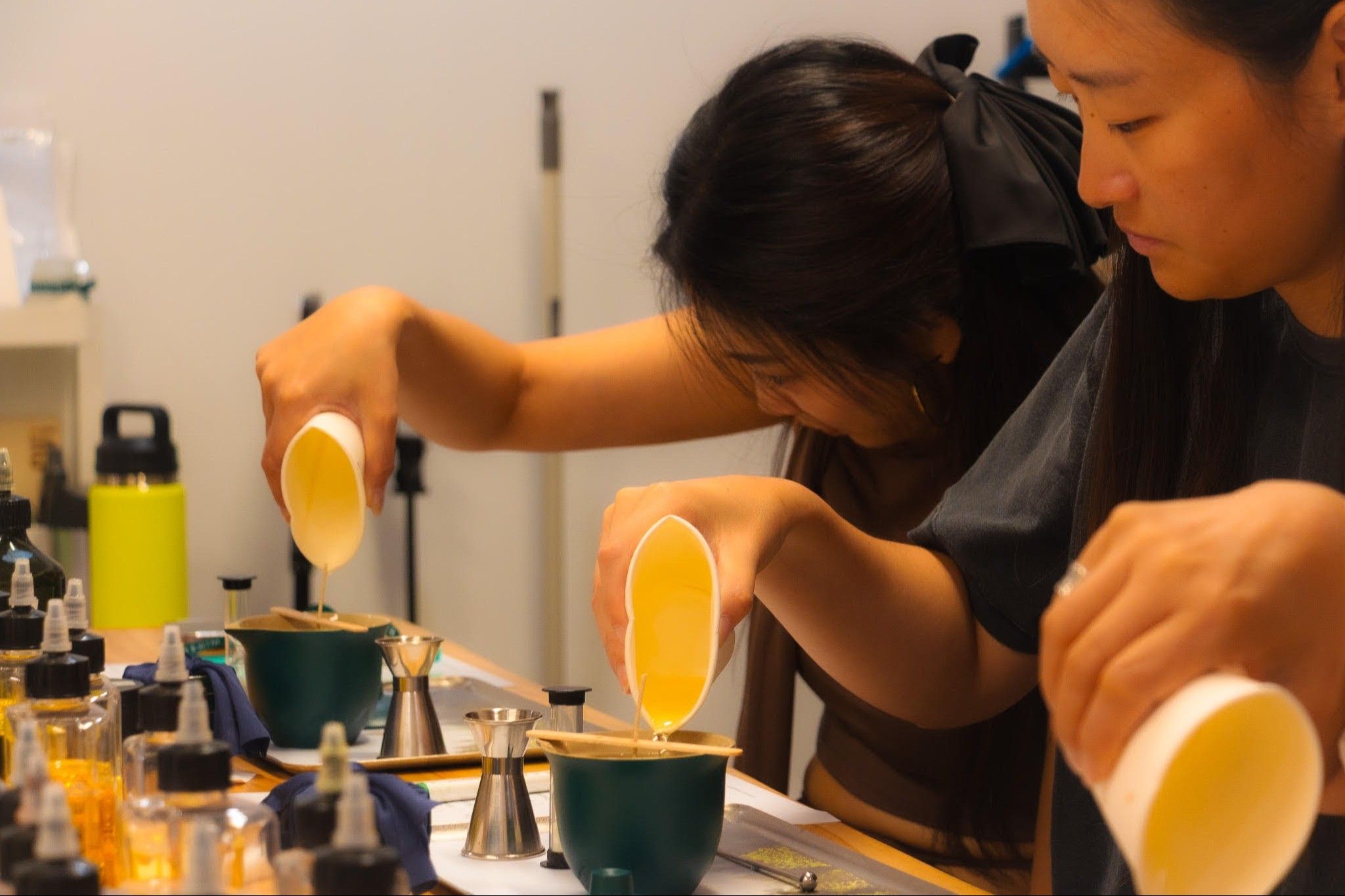 Three people pouring liquid from white cups into green containers on a table with various bottles and tools.