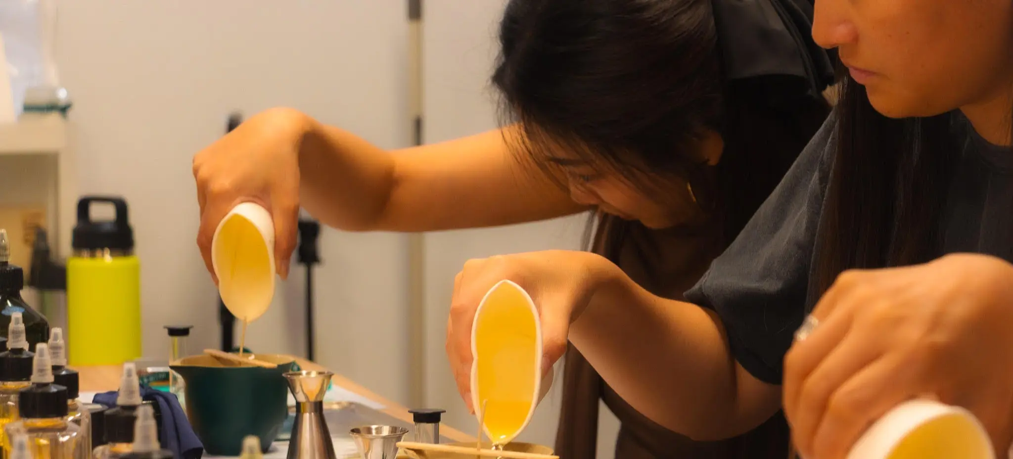 Three people pouring liquid from white cups into green containers on a table with various bottles and tools.