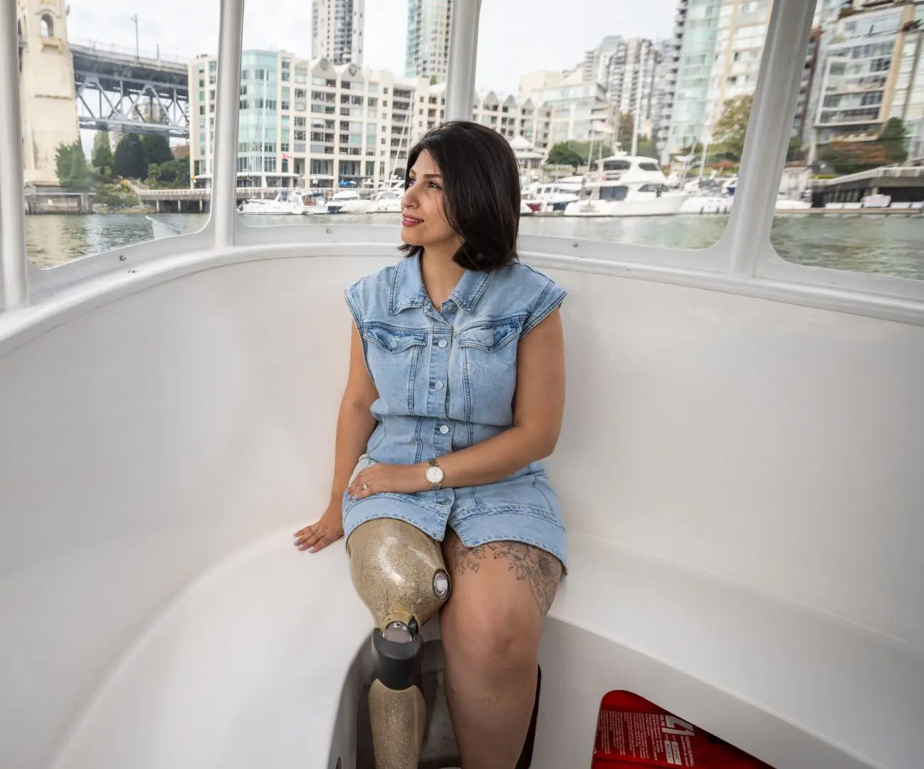 A woman with a prosthetic leg sits relaxed inside a bright, windowed ferry boat, enjoying a scenic ride across the water with the city skyline and a bridge in the background.