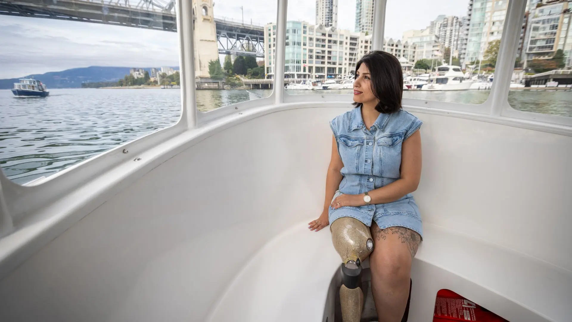 A woman with a prosthetic leg sits relaxed inside a bright, windowed ferry boat, enjoying a scenic ride across the water with the city skyline and a bridge in the background.