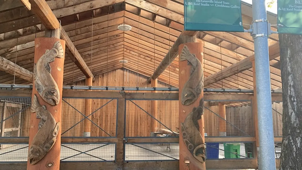 Wooden structure with a peaked roof and carved wooden poles featuring fish designs inside a fenced area.