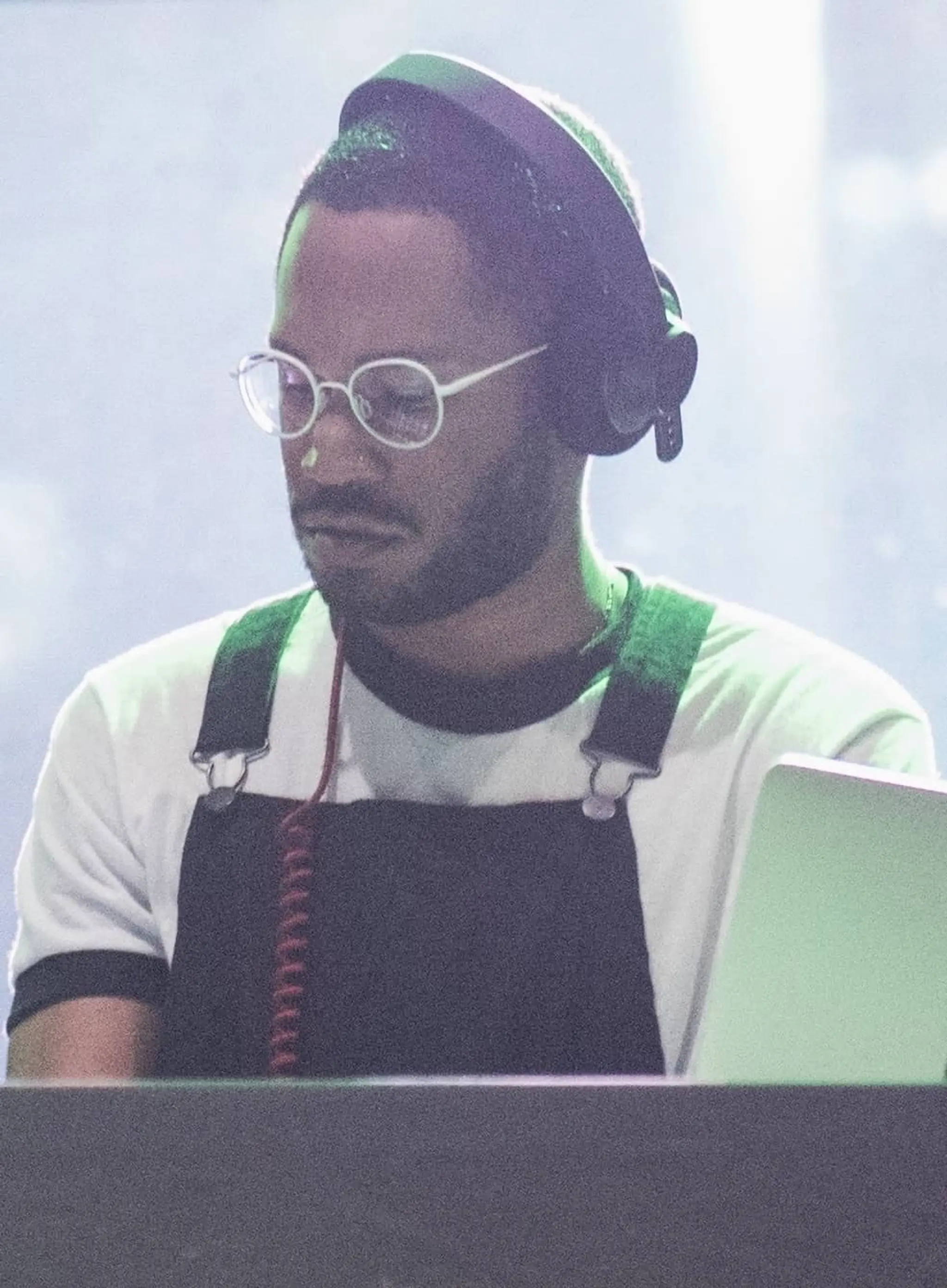 Man wearing glasses and headphones, focused on a laptop, dressed in a white shirt with black overalls.