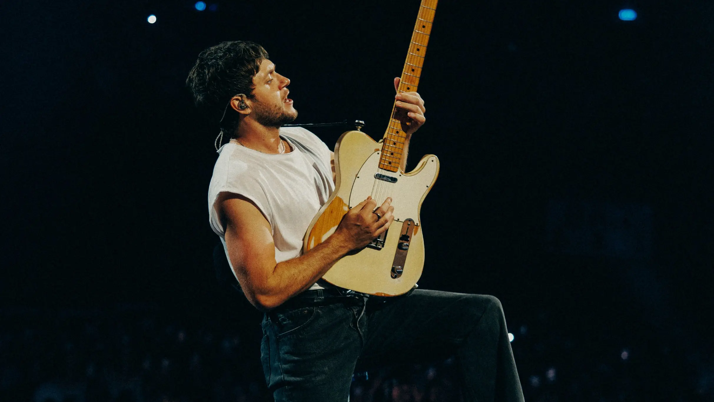 A person playing an electric guitar on stage wearing a white sleeveless shirt and dark pants against a dark background.